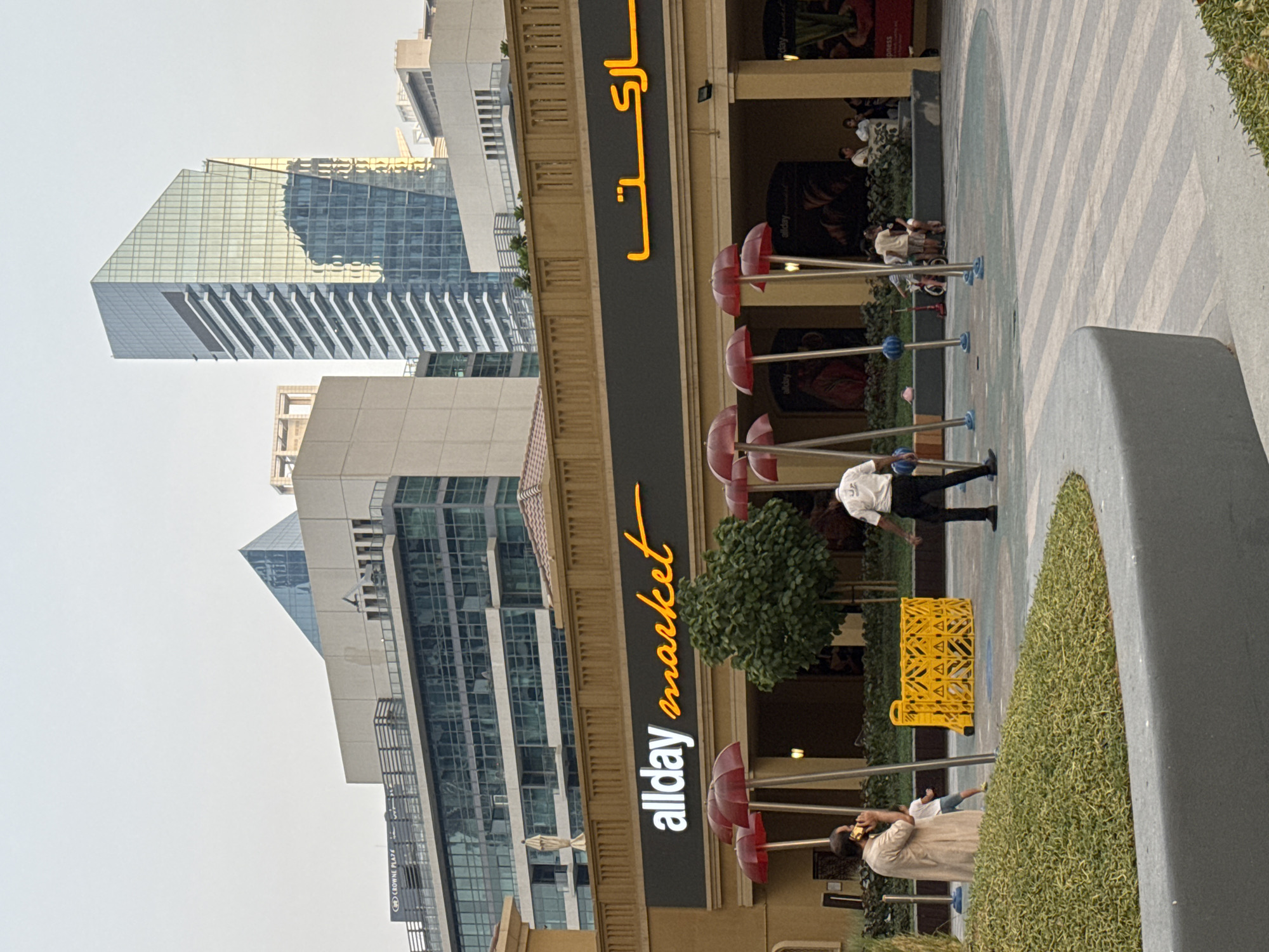 The image captures a street-level view of "allday market" in Dubai, United Arab Emirates, with modern architecture in the background. In the foreground, a tan-colored building with a dark awning prominently displays the "allday market" logo in orange script and white sans-serif font. Below the awning, a series of red umbrella-like structures on poles are visible, and a small tree stands between two of them. On the left, a person in a tan robe is on a phone call, and a child in a white shirt and blue shorts is nearby. To the right, a person in a white uniform and black pants walks away from the camera. In the middle ground, a yellow plastic barrier is set up, and further back, a group of people are gathered near the entrance of the market. The background features several contemporary buildings, including a prominent skyscraper with a blue glass facade and angular design, reflecting the sky. The sky is a pale, hazy blue, suggesting it might be late afternoon or early morning. The ground is paved with a patterned surface.