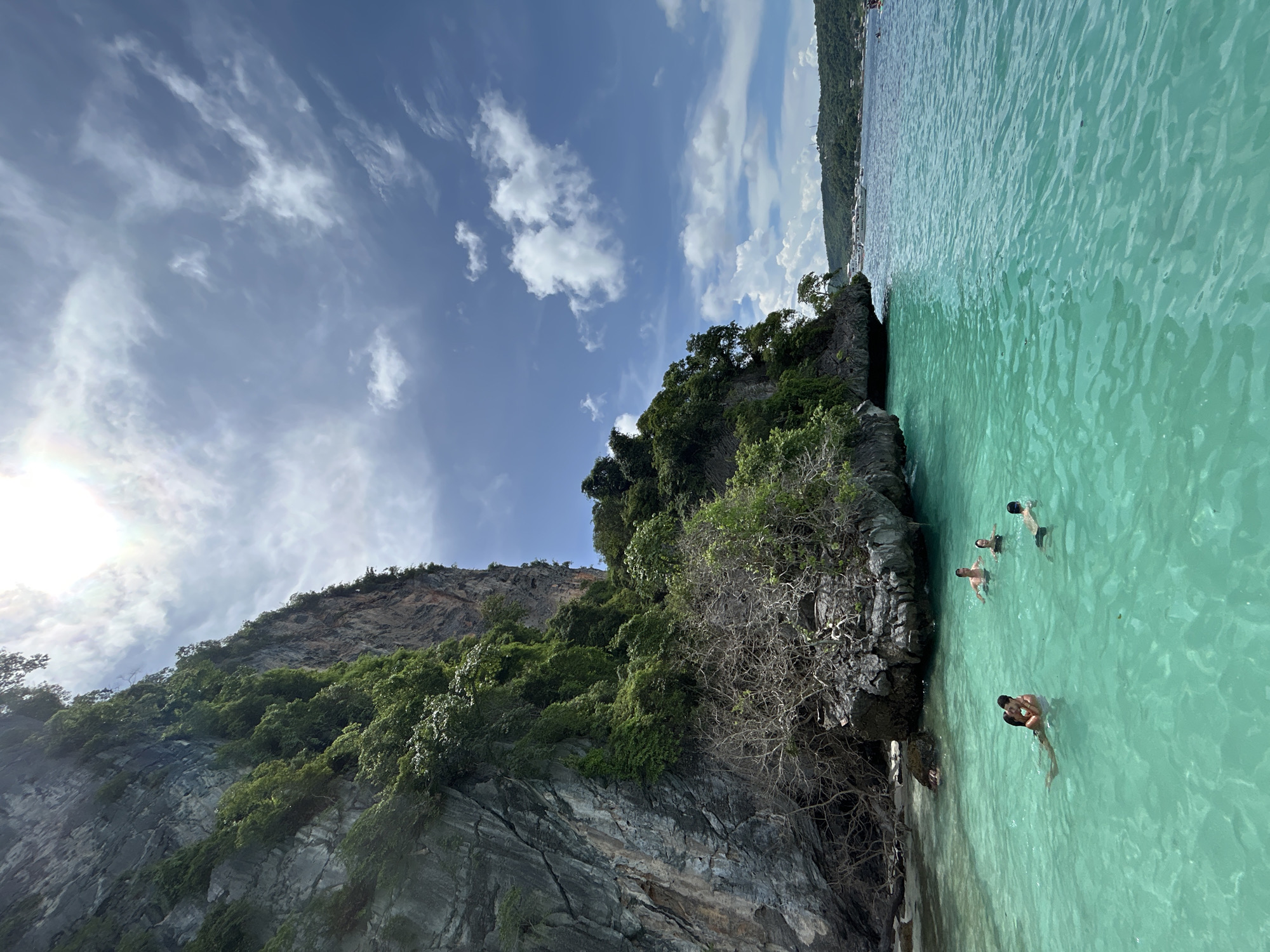 A vertical, eye-level shot captures a tropical scene with a towering limestone cliff on the left and vibrant turquoise water on the right. Several people are enjoying the water, with one couple in the foreground embracing while another individual swims nearby. In the background, the water extends to a hazy horizon with lush green hills and a few boats. The sky is a bright blue, dotted with scattered white clouds, and the sun is visible, casting a bright glare. The limestone cliff is densely covered with vegetation, with dry, thorny branches protruding from its base. The water is crystal clear, revealing the sandy bottom in some areas. The scene appears to be from the islands off the coast of Ko Lanta, Thailand.
