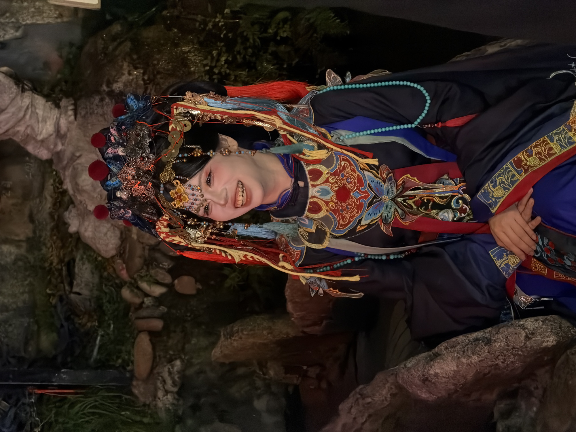 The image shows a young East Asian woman dressed in elaborate, traditional Chinese opera attire. She has a pale complexion, dark hair styled in an updo with numerous ornaments, and her face is painted with heavy makeup, including bright red blush and eyeliner. She is smiling broadly, revealing her teeth. Her costume is a deep navy blue with intricate gold, red, and blue embroidery featuring floral and butterfly motifs. She wears a headdress adorned with colorful feathers, beaded chains, and what appear to be small red pom-poms. The background is dark and appears to be a rocky, naturalistic setting, possibly a garden or a set designed to resemble one, with some greenery and stones visible. The lighting is dim, with the woman being the primary light source. The woman is seated or resting against a large, textured rock on the left side of the frame, with her hands clasped in her lap. The overall impression is that of a performer in costume, possibly backstage or in a scenic area. The location is identified as Yuzhong District, China.