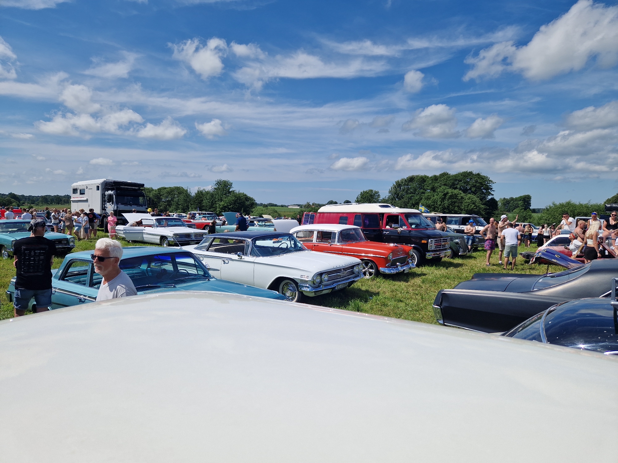 The image depicts an outdoor gathering, likely a car show, taking place in a grassy field in Edenberga, Sweden. The sky is bright blue with scattered white clouds. In the foreground, a portion of a white car's hood is visible, obscuring the lower part of the scene.
Behind the foreground car, several vintage and classic cars are parked, including a light blue four-door sedan with a silver roof, a white two-door sedan with a black roof, and a bright orange two-door sedan. A man with gray hair and sunglasses stands near the light blue sedan, appearing to be interacting with someone off-camera or observing the vehicles.
Further in the background, more vehicles are present, including a large white camper truck with a black roof rack. A group of people are milling about, some standing in small clusters and others walking between the cars. The atmosphere appears relaxed and social. Trees and foliage form a green backdrop beyond the field of cars and people. The event is taking place on a sunny day.