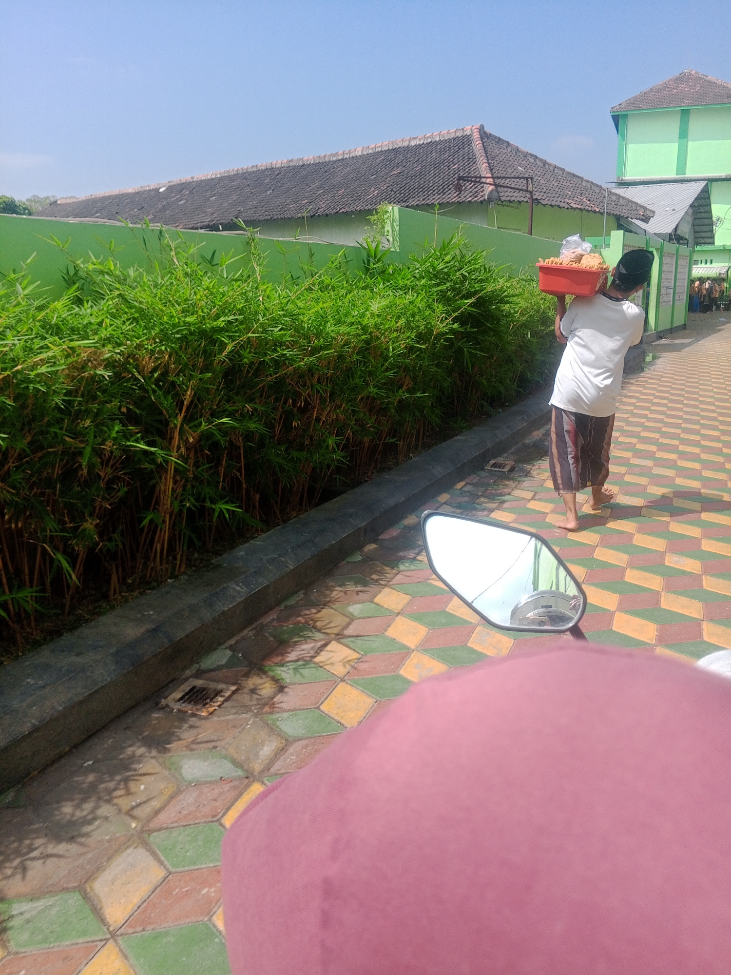 The image shows a person carrying a red plastic container filled with food items on their right shoulder. The person is wearing a white t-shirt and striped pants, with a black cap on their head. They are walking on a paved pathway with a geometric pattern of green, red, and yellow tiles. To the left of the pathway is a lush green bamboo hedge, bordered by a dark gray concrete structure. In the background, there are two buildings; one with a dark tiled roof and a light green wall, and another larger building that is predominantly light green with some gray elements. The sky is clear and blue. The scene appears to be an outdoor area, possibly a school or a religious institution, given the architectural style and the attire of the person. The overall impression is that of a warm, sunny day in an urban or semi-urban environment in Ponorogo, Indonesia.