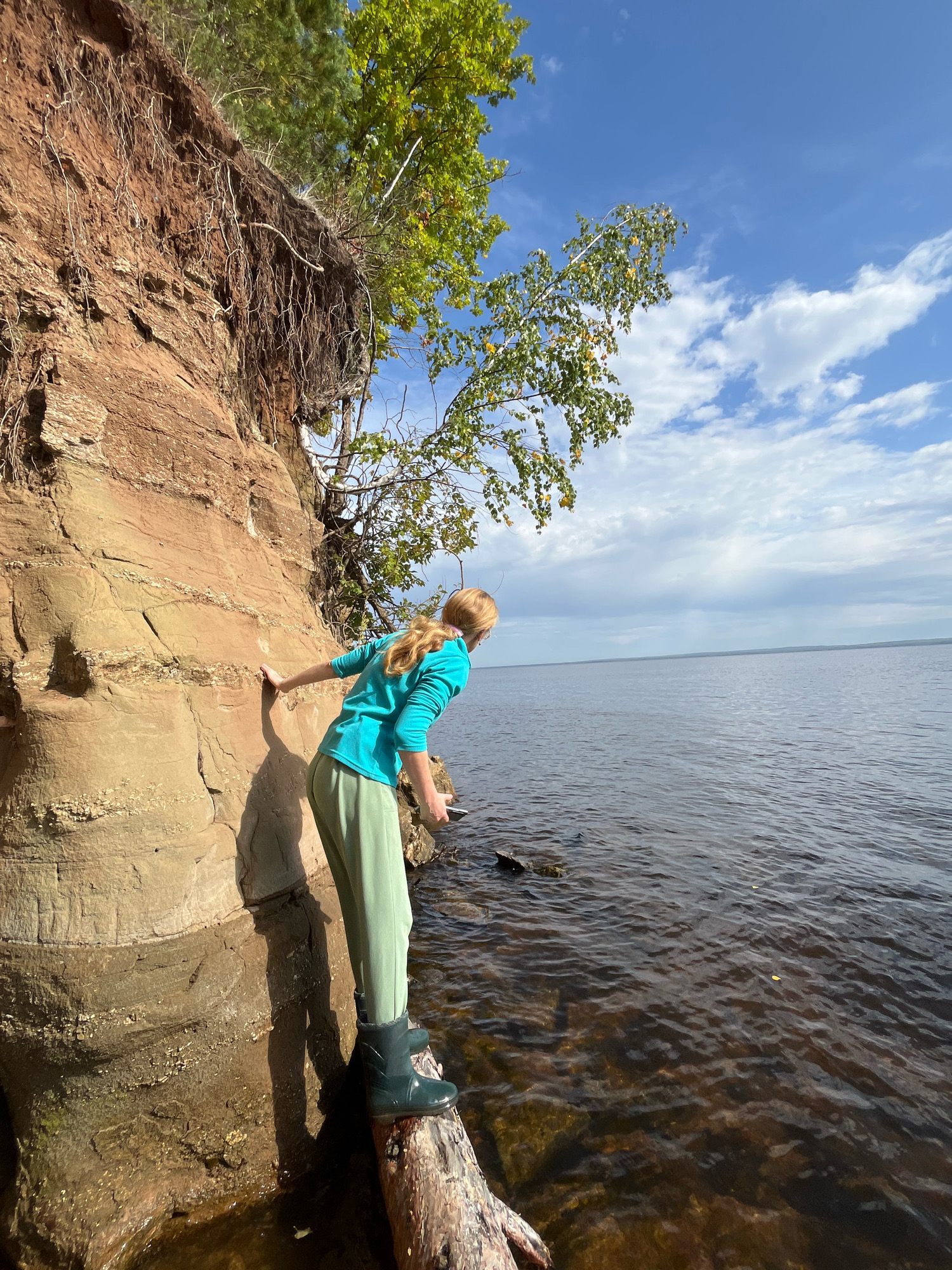 A young woman with blonde hair pulled back stands on a fallen log at the edge of a cliff overlooking a body of water. She is wearing a teal long-sleeved shirt and light green pants, with black rain boots. Her right hand is resting on the reddish-brown cliff face. The cliff is eroded and shows distinct layers of sediment. Trees with green and yellowing leaves grow from the top of the cliff. The water is dark and appears calm, with ripples on the surface. The sky is blue with scattered white clouds. The scene suggests a natural landscape, possibly in a park or wilderness area. The location is identified as the Chaikovsky Reservoir, a man-made lake on the Kama River near the city of Chaikovsky, Russia.