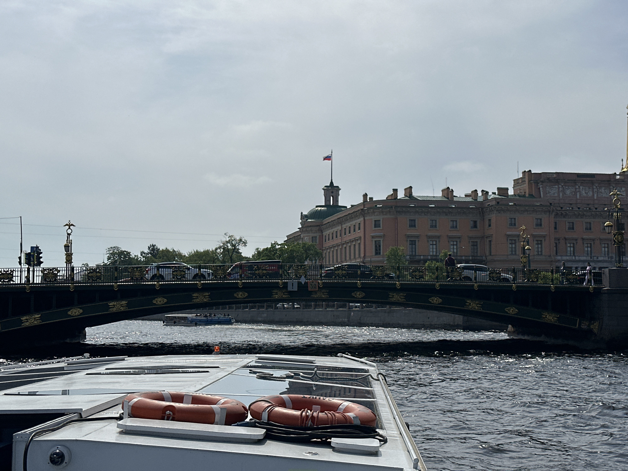 The photograph captures a scene in Saint Petersburg, Russia, featuring a boat navigating a waterway beneath a bridge. Two orange life buoys are prominently displayed on the deck of the boat. The bridge, constructed with dark green and gold ornate ironwork, carries vehicular and pedestrian traffic. Behind the bridge, a large, multi-story building with a domed tower topped by a Russian flag is visible. The architecture suggests a historical or governmental structure. The sky is overcast, and the water is moderately choppy, reflecting the ambient light.