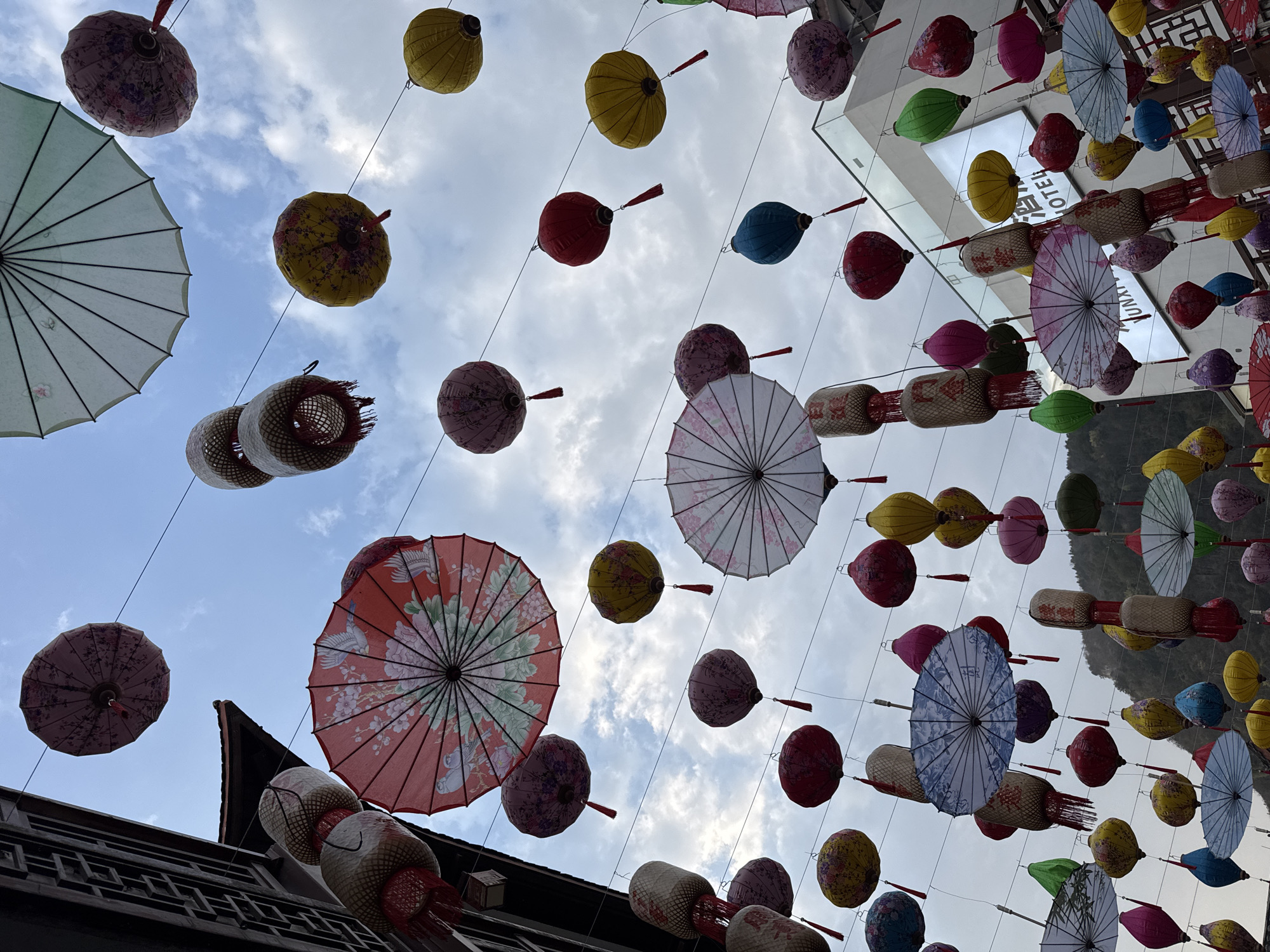 The image presents an upward-angled view of a street or alleyway in Wulingyuan, China, densely adorned with overhead decorations against a partly cloudy sky. Numerous traditional Chinese lanterns and decorative umbrellas are suspended by thin, dark wires stretching across the frame.
The lanterns exhibit various shapes and colors: spherical lanterns in red, yellow, pink, green, blue, and purple, often decorated with floral motifs and terminated with tassels. Elongated cylindrical lanterns, primarily in beige woven material, also feature red tassels. Some lanterns are gourd or teardrop-shaped.
Interspersed among the lanterns are open, circular paper or silk umbrellas. These umbrellas are patterned with intricate designs including cherry blossoms, peonies, birds such as cranes and sparrows, and abstract landscapes, rendered in colors like red, white, pink, blue, and light green.
On the left side of the frame, a portion of a traditional Chinese building is visible, characterized by dark wooden lattice work, window frames, and a curved, dark tiled roof. To the right and in the mid-ground, a modern multi-story building with white facades and dark-framed glass panels is present. Partial white signage for "HOTEL" is visible on its exterior. In the distant background, lush green mountains rise, framing the urban setting. No individuals are depicted within the scene.