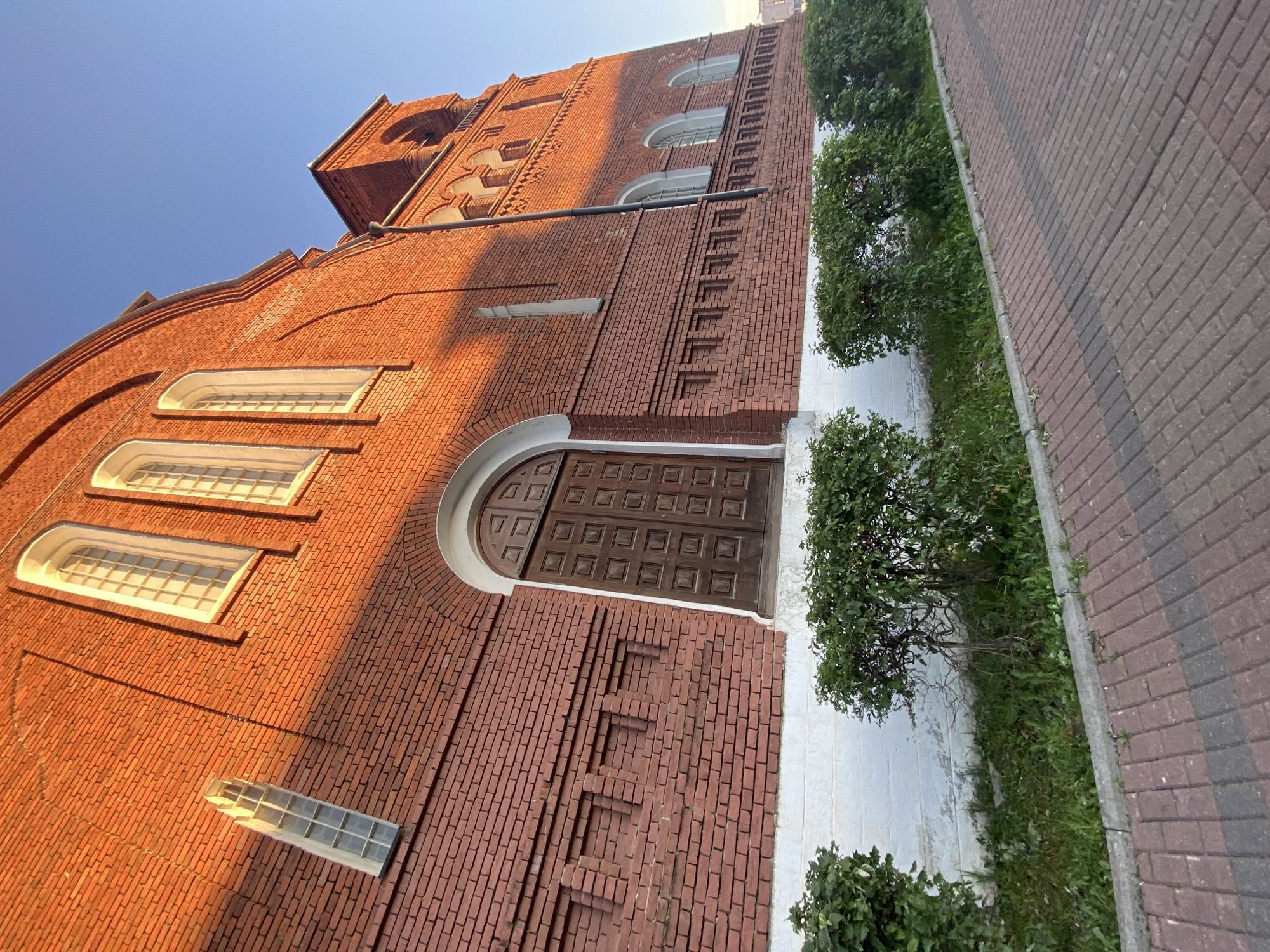 The image shows the exterior of a red brick building with a clear blue sky in the background. The building features arched windows with white frames and multiple panes of glass. A large, dark wooden double door with a decorative pattern is visible, set within a rounded archway. The brickwork on the building has a textured, decorative pattern in places. A white-painted base runs along the bottom of the brick wall.
To the right of the building, there is a paved walkway made of interlocking bricks in shades of red and gray. A narrow strip of green grass and small bushes borders the walkway, separating it from the building. The lighting suggests it is either early morning or late afternoon, with long shadows cast across the brickwork. There is no visible text in the image. The scene depicts a quiet exterior of a building, possibly a historical or religious structure, in a well-maintained outdoor setting. The image is oriented vertically, with the sky at the top left and the ground at the bottom right.