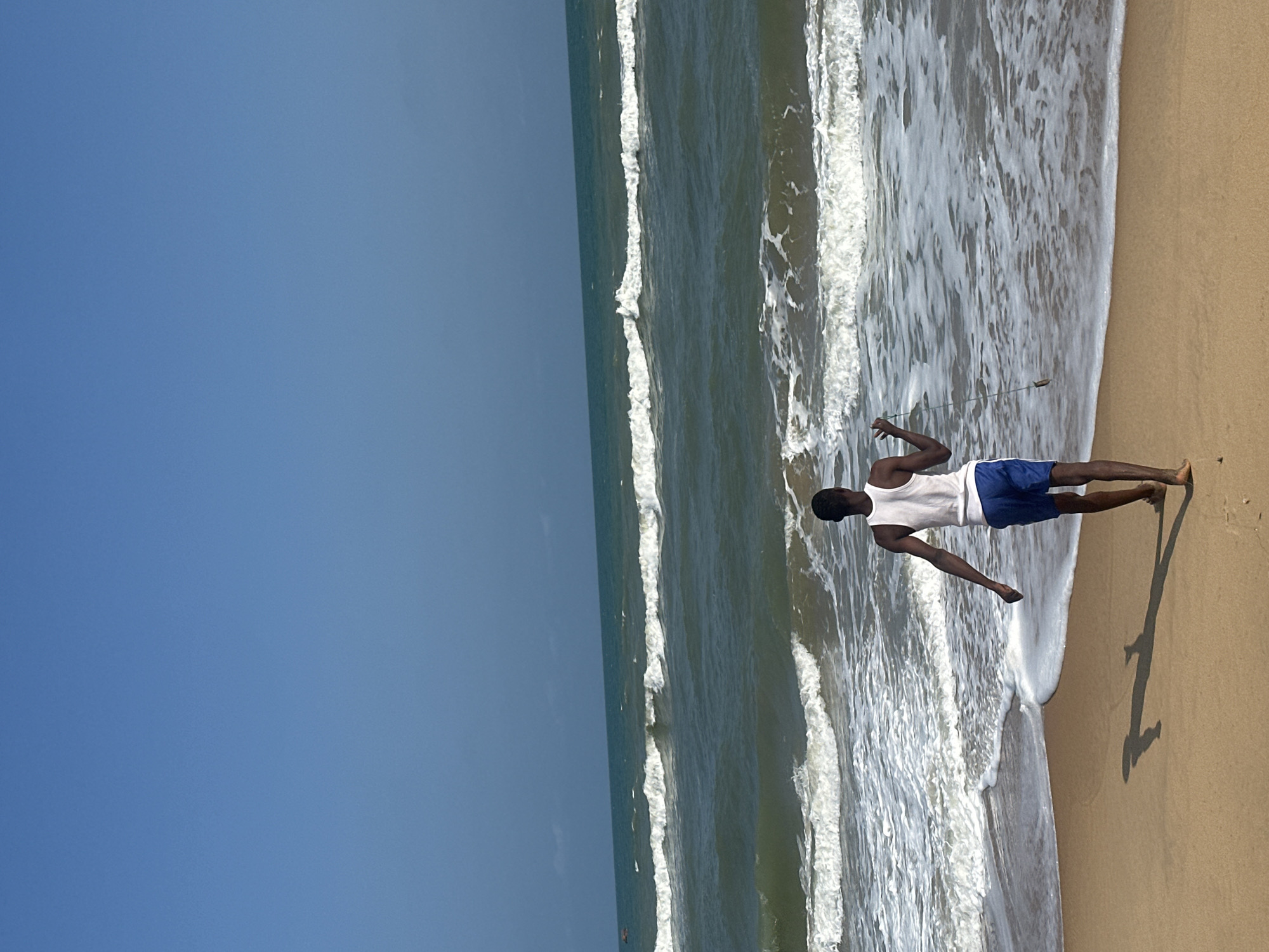 A young Black man stands on a sandy beach, with his body facing the ocean. He is wearing a white tank top and blue shorts. The man is casting a fishing line into the water, with a small weight at the end of the line. Waves are crashing on the shore, and the water is a deep green color. The sky is a clear blue, and the sun is high in the sky, suggesting it is daytime. The man's shadow is cast long on the sand, indicating the sun is somewhat low in the sky, likely late afternoon or early morning. The scene appears to be a peaceful, relaxing day at the beach. The location is identified as Badagry, Nigeria. There is no visible text in the image.