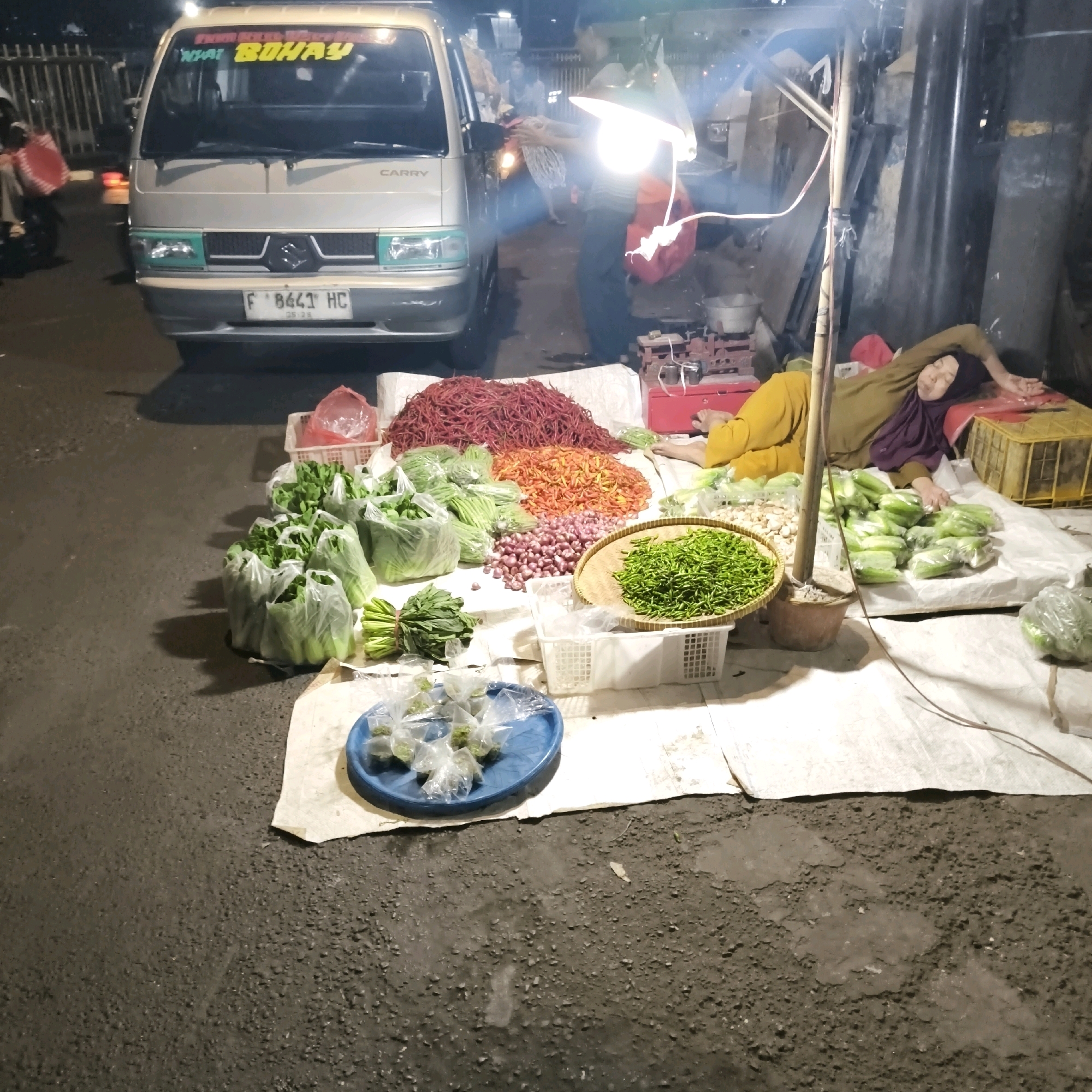 A woman wearing a purple hijab and a mustard-colored outfit lies on her side on a white tarp, resting her head on her arm. She appears to be asleep. The tarp is spread on asphalt, likely a roadside stall in South Jakarta, Indonesia. A large pile of red chilies is visible behind her, alongside heaps of orange chilies and red onions. To the left of the chilies, several plastic bags filled with leafy green vegetables are stacked. In front of the vegetables, a woven basket holds a pile of green chilies. Another blue plate with small, individually wrapped bundles of herbs or spices sits on the tarp near the foreground. A silver van with the word "CARRY" on its back is parked in the background, and a bright lamp hangs overhead, illuminating the scene. A red scale and a red chest are also visible, suggesting a market setting.