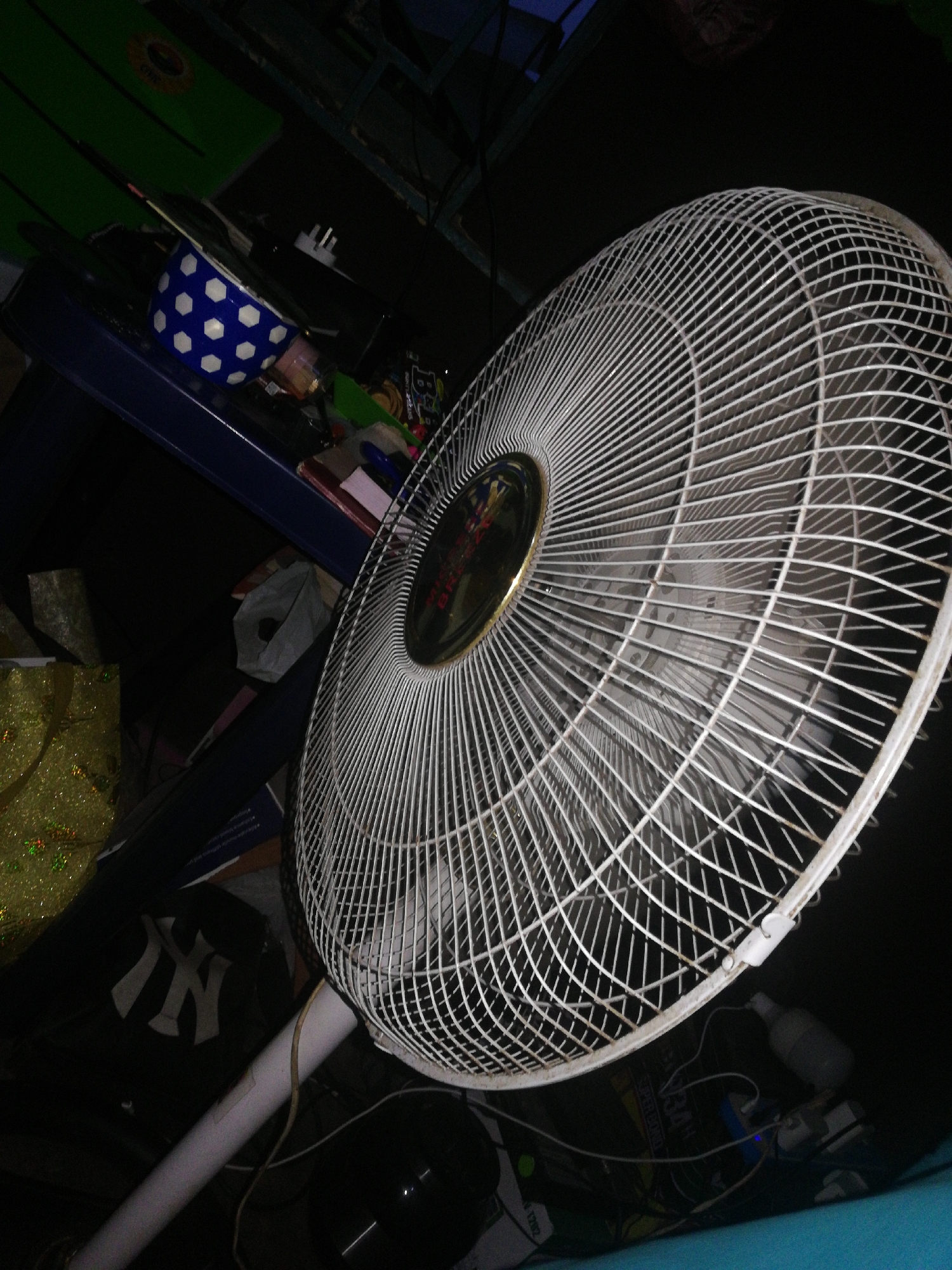 The image shows a white standing fan with a round metal safety grill. The fan is positioned prominently in the foreground. Behind the fan, there is a dark and cluttered background. A blue polka-dotted bowl sits on a blue surface. To the left, there is a black item that has the NY logo. Further back, a green surface is visible. Wires and various objects are scattered around the base of the fan. The lighting appears dim.