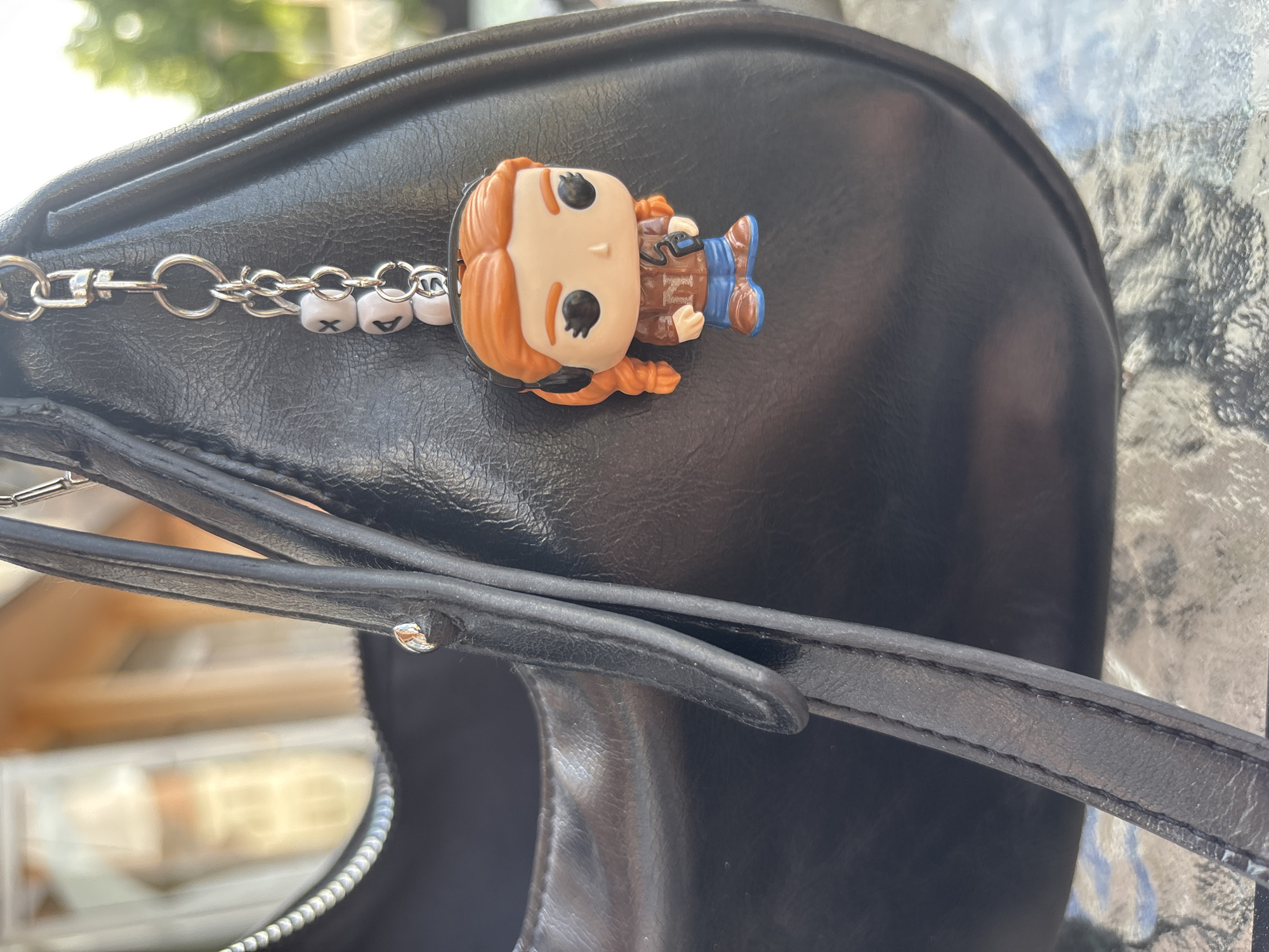 The image shows a close-up of a black leather purse resting on a textured surface, possibly a glass table. A Funko Pop figurine of a red-haired character, likely a female, is attached to the purse via a chain with lettered beads. The purse strap is visible in the foreground. The background is blurred, obscuring the surroundings. Without further information, the exact location in Garmen, Bulgaria, cannot be determined from the image alone. The scene depicts a personal item with a decorative element in an outdoor setting.