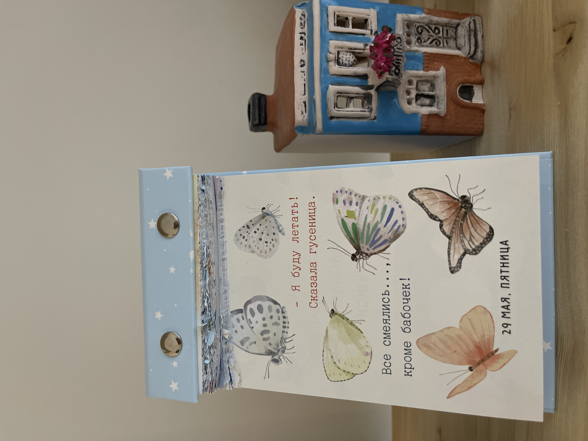 The image shows a close-up of a desk calendar next to a ceramic house figurine. The calendar is open to a page with illustrations of butterflies and Russian text, indicating the date as "29 May, Friday." The ceramic house figurine is blue and brown, with small details like windows and a balcony with flowers. The background is a plain wall.