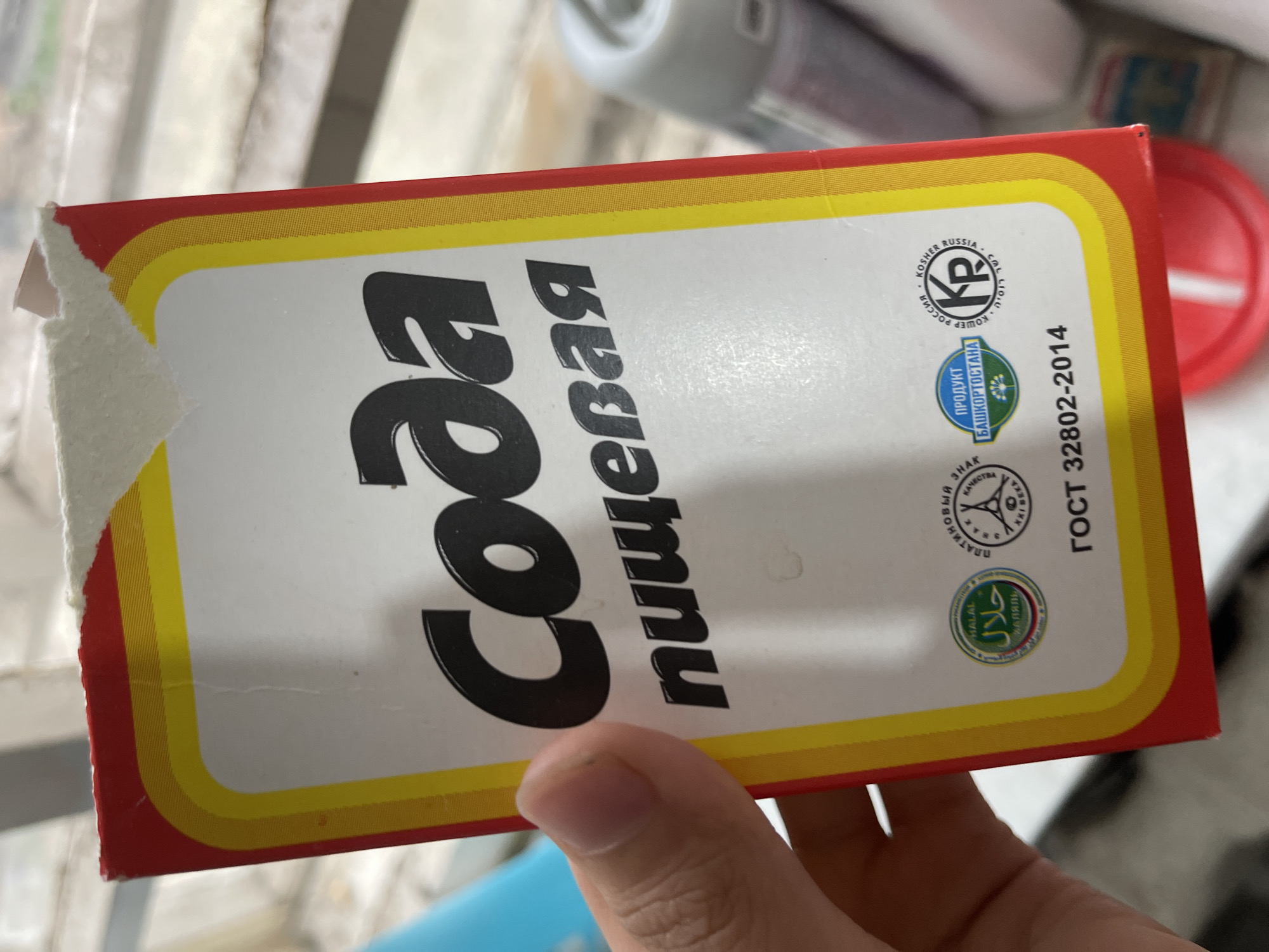 A hand is holding a red and yellow box of "Soda Pishchevaya" (baking soda) in what appears to be an indoor setting, possibly a kitchen or pantry. The box has Cyrillic text and several certification logos, including "Halal," a blue and green logo indicating "Product of Bashkortostan," and a "Kosher Russia" symbol. The background is out of focus but suggests shelves with containers and possibly a window with light filtering through. The image is shot from a slightly upward angle.