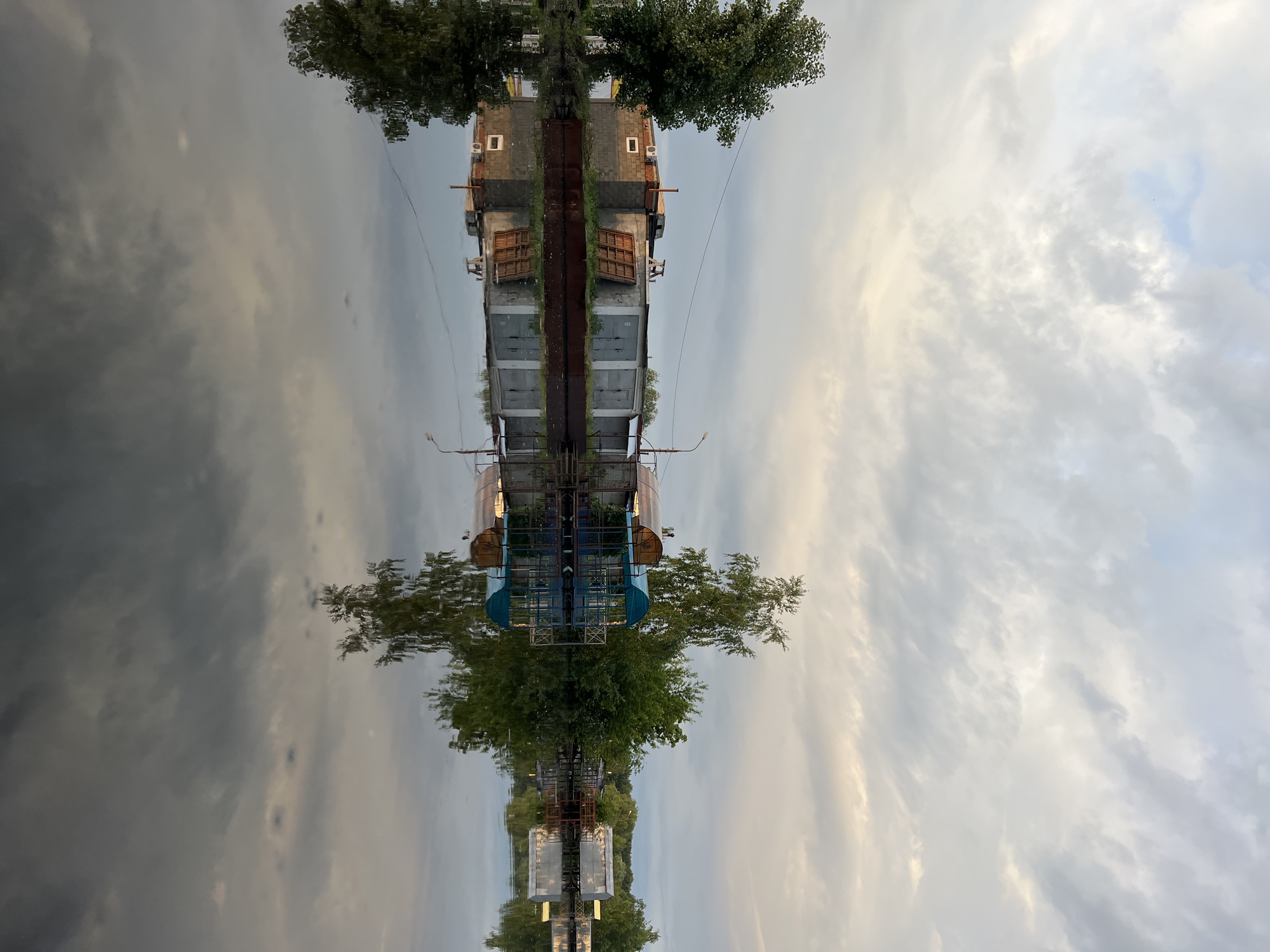 The image displays a serene waterside scene, captured and presented rotated 90 degrees clockwise. If viewed correctly (rotated 90 degrees counter-clockwise), the upper half depicts the actual landscape, while the lower half shows its reflection in the water.
The primary subject is a large, multi-level man-made structure, possibly a bridge or a repurposed industrial platform, spanning across or situated near a body of water. This structure appears constructed from concrete and metal, featuring various components. One section resembles a building with a gabled roof, small rectangular windows, and what might be air conditioning units. Another part exhibits more open metalwork, railings, and supports, with several light poles or street lamps integrated. Lush green trees with dense foliage are prominent, growing both along the water's edge and seemingly from parts of the structure itself. Above, the sky is largely covered by soft, diffused clouds, ranging from light grey to white, with subtle warm hues. The water below acts as a mirror, faithfully reflecting the sky, trees, and the entire structure, with slight ripples creating gentle distortions in the reflections.
No human activity or specific event is taking place; the scene is static and peaceful.
Notable details include the soft, diffused lighting, which suggests either early morning or late afternoon/evening. The weather appears overcast or partly cloudy. The striking symmetry created by the reflection in the water is a key compositional element. The combination of industrial architecture and natural elements aligns with a waterside location in Chaikovsky, Russia. The overall mood is tranquil and contemplative.
There is no visible text in the image.