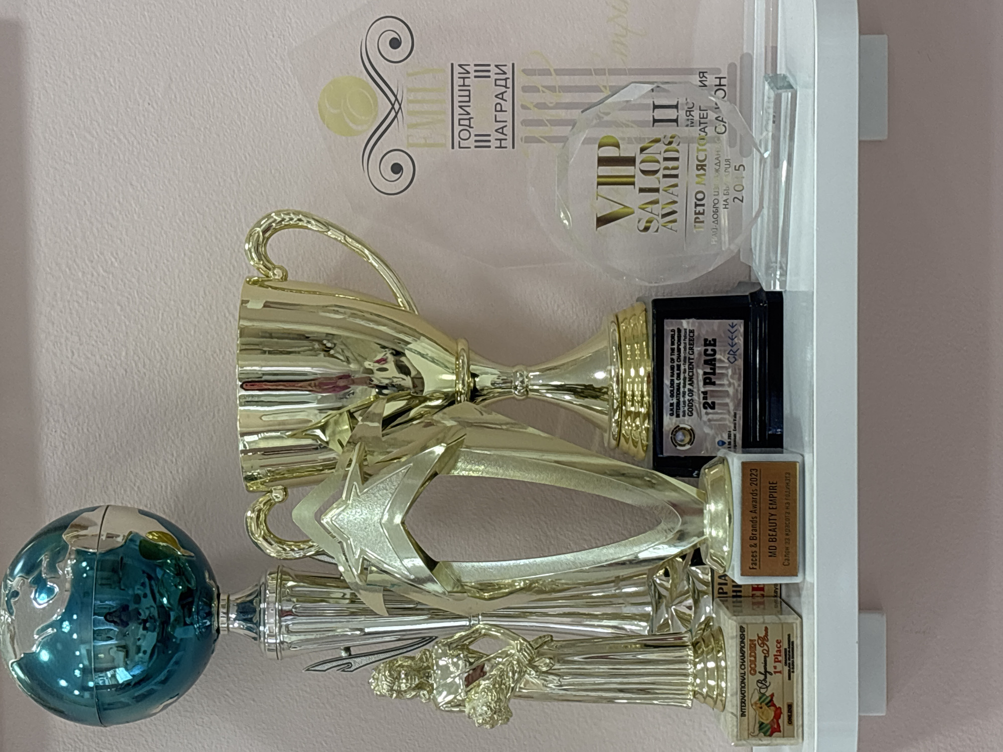 The image shows a collection of awards and trophies displayed on a white shelf against a light pink wall. The trophies vary in size and design, with some appearing gold and silver. There is also a blue decorative globe on the left.
Several of the awards have inscriptions. One reads "Faces & Brands Awards 2023, MD BEAUTY EMPIRE, Салон за красота ка годината" indicating that the awards are for beauty salons and are specific to the year 2023. Another reads "VIP SALON AWARDS II", implying a salon-specific award. Another is for a "INTERNATIONAL CHAMPIONSHIP GOLDEN" award, placing first. Also shown is a second place award for "GJHR-GOLDEN HAND OF THE WORLD INTERNATIONAL ONLINE CHAMPIONSHIP".
The text "ТРЕТО МЯСТО КАТЕГИЯ НАЙ-ДОБРО ИЗВЕЖДАНЕ СА ОН НА БЪ АРИЯ 2015" suggests a third place award for the best service salon in Bulgaria in 2015. The phrase "Салон за красота ка годината" suggests that the salon is located in the city of Sofia, Bulgaria.
