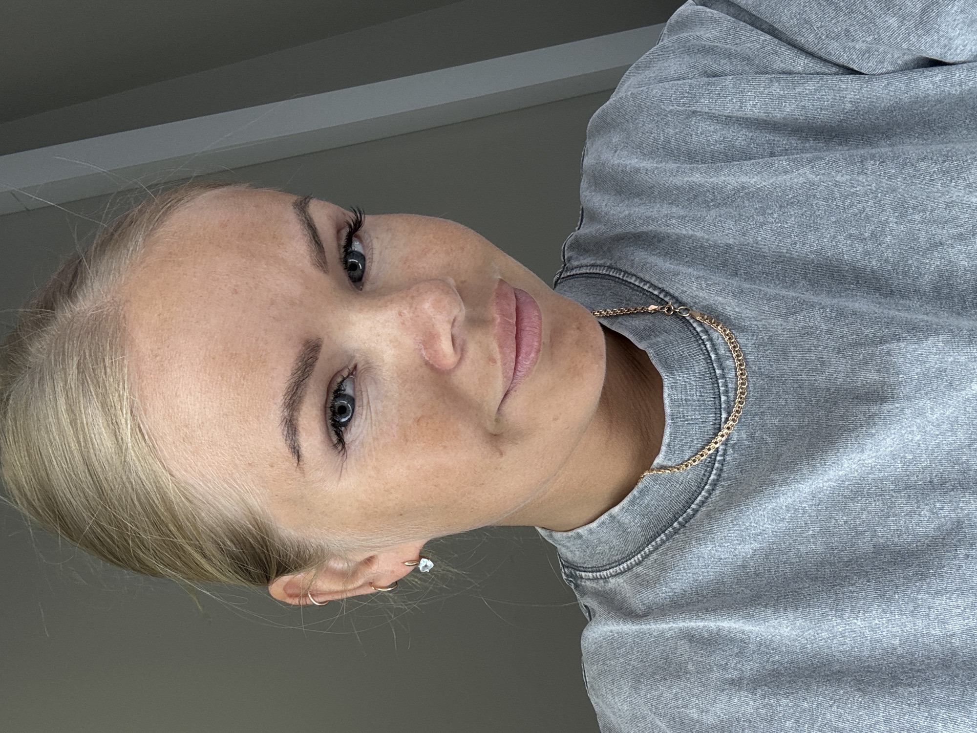 A close-up, angled portrait shows a woman with blonde hair pulled back. She has fair skin with freckles and visible eyelashes. She is wearing a grey sweatshirt with a gold chain. Her left ear has two gold hoop earrings and one gemstone stud earring. The background is a light grey color. The image appears to be taken indoors. The location is Unknown, Unknown.