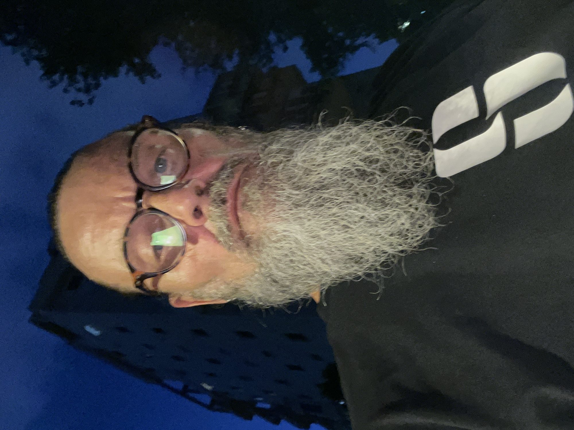 The media file is a self-portrait of a man with a long, gray beard and glasses. He is wearing a black t-shirt with a white logo consisting of two curved shapes that form a stylized "S". The man appears to be outdoors at night, with a dark blue sky visible behind him. In the background, there is a building with lights on, suggesting an urban setting. The lighting suggests it is evening or night. The man's face is close to the camera, and his expression is neutral. The overall mood of the image is casual and personal, typical of a selfie. The provided location context is Netanya, Israel.