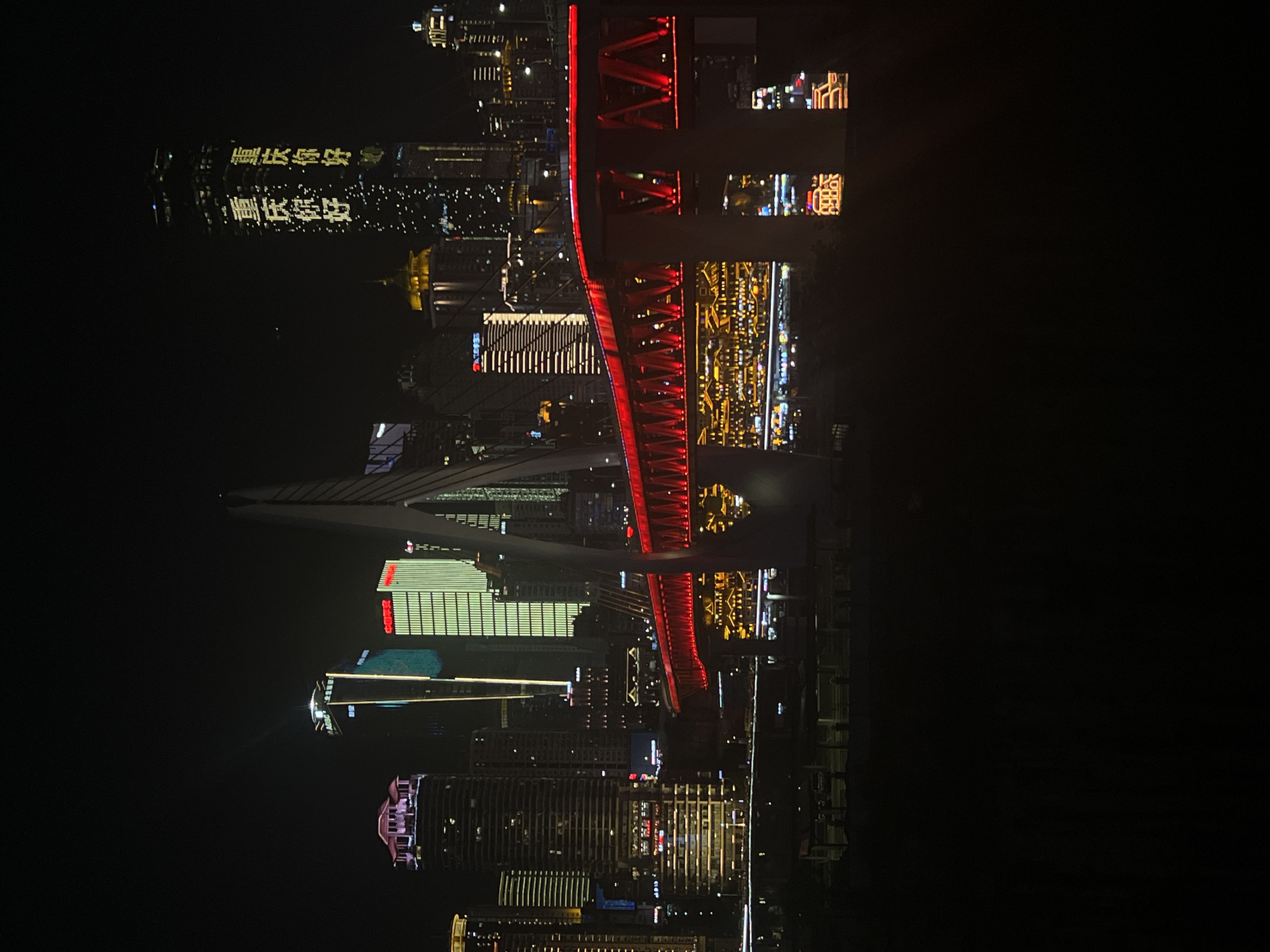 The image shows a nighttime cityscape with a prominent illuminated red bridge spanning across a body of water. Tall buildings with bright lights are visible in the background. On one of the buildings, large yellow characters, which read "重庆你好" (Chongqing Hello), are displayed. The scene is dark, indicating it is night. The location is identified as Jiangbeicheng Subdistrict, Chongqing, China.