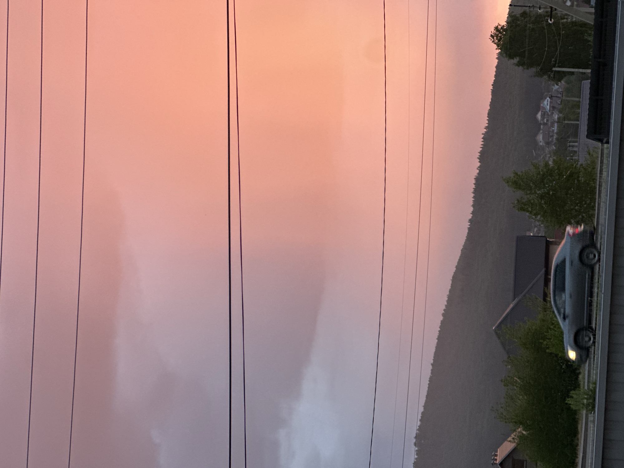 The image, oriented horizontally, captures a twilight or dawn scene featuring a colorful sky, a forested hillside, and elements of a built environment.
The upper portion of the frame is dominated by the sky, which transitions from a soft pink and orange at the top to a lighter, more muted pink and grey towards the bottom, indicating either sunrise or sunset. Multiple dark utility lines or power cables stretch horizontally across the entire width of the sky.
In the middle ground, a densely forested hill or mountain range slopes upwards from left to right, its treeline silhouetted against the sky. Scattered among the trees on the hillside are a few houses or buildings with visible roofs.
The foreground on the right side of the image features a dark, elongated structure, possibly a building or part of an overpass. On a road or elevated platform next to this structure, a silver or light-colored car is visible, its front end facing right. The car's headlights appear to be on, emitting a warm glow. Below the road/platform, more trees and bushes are present, partially obscuring other structures or ground features.
No people are visible. No discernible text is present in the image. The overall atmosphere conveyed is calm and somewhat moody due to the subdued lighting and sky colors.