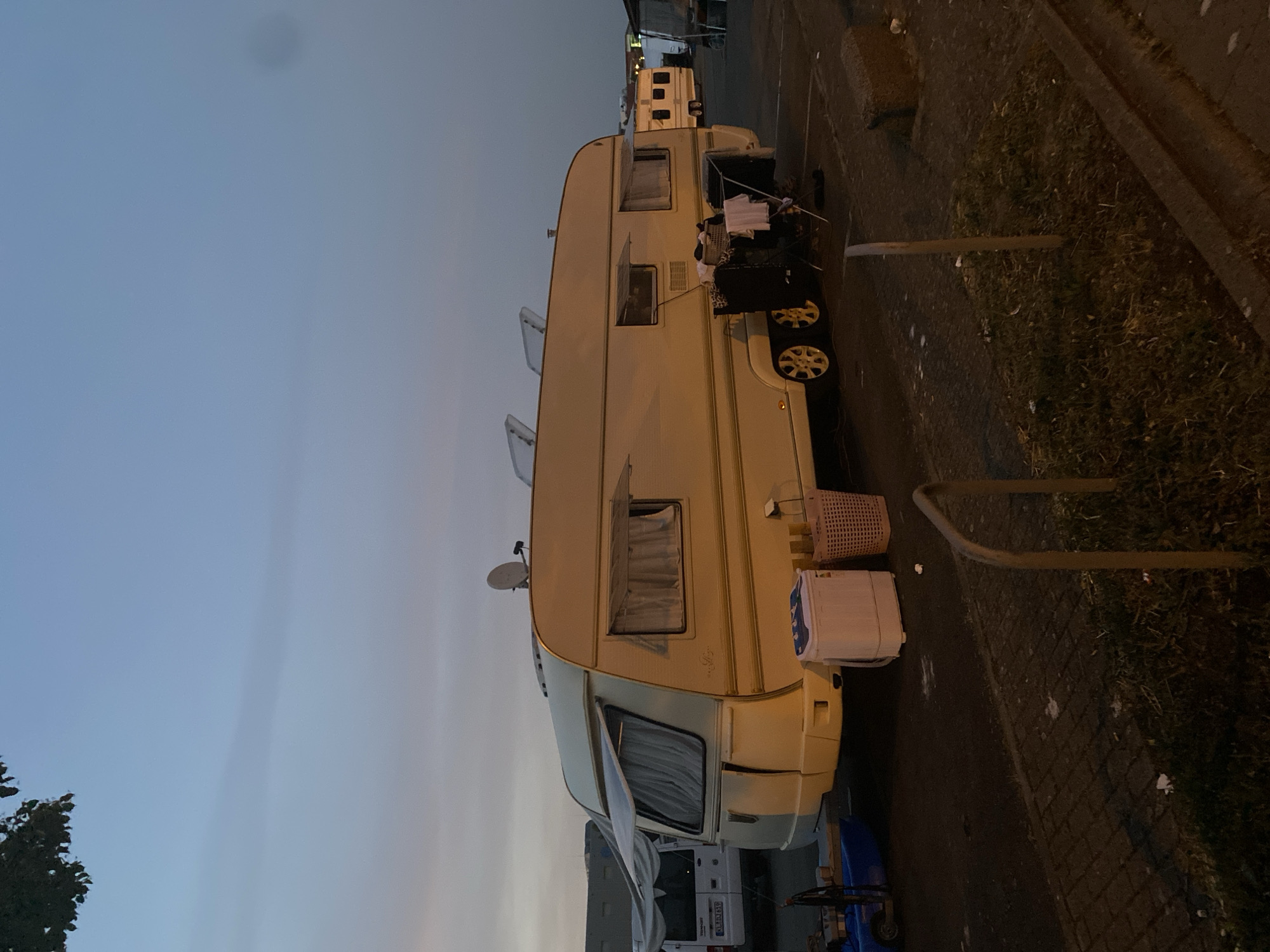 A horizontal outdoor shot shows a white caravan parked on a paved street in Landau in der Pfalz, Germany. The sky is a gradient of light blue to gray, indicating dusk or dawn. The caravan, bearing a satellite dish on its roof, has windows with curtains. A white washing machine and a pink plastic basket sit next to the caravan. A clothesline extends from a window with clothing hung to dry. A bicycle rack is visible near the sidewalk. In the background, more caravans are parked along the street. Debris is scattered on the ground. License plate of one of the vehicles has a German plate with "LD-FN 519".