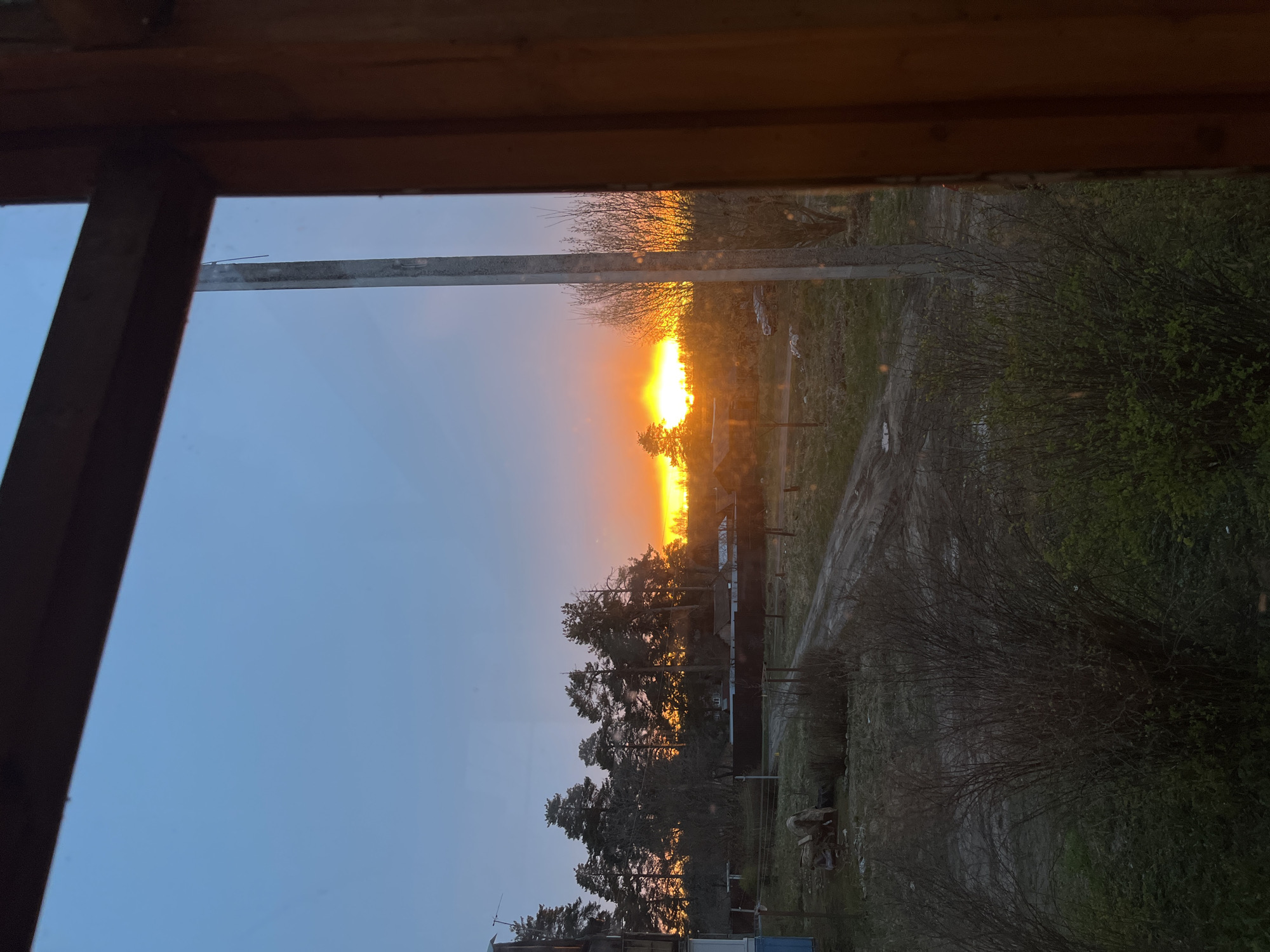 View through a window frame of an orange sunset in Раздолье, Russia. The sky transitions from blue to orange near the horizon, where the sun is visible through trees and structures. A dirt road is seen to the right of the trees, along with sparse vegetation.