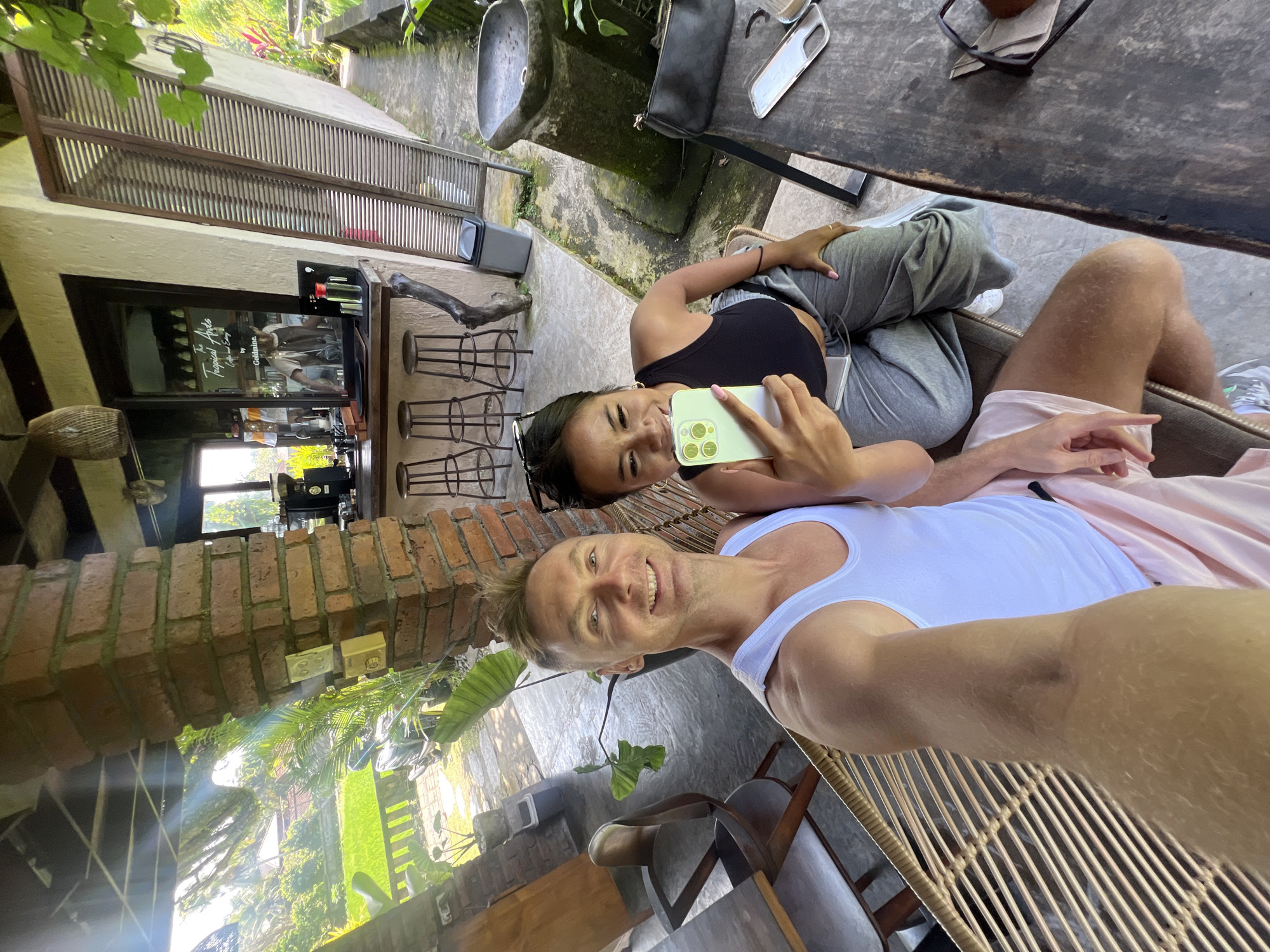 In Ubud District, Indonesia, two individuals are seated on rattan chairs on a patio, appearing to be at an outdoor cafe or restaurant. The man, wearing a white tank top and pink shorts, is taking a selfie with his phone. Beside him, a woman in a black crop top and grey sweatpants holds a smartphone, looking at the camera and smiling. Behind them, a brick pillar supports a roof structure. A bar area with stools and coffee equipment is visible in the background. The patio opens onto a lush green landscape with rice paddies and tropical plants, suggesting a rural or semi-rural setting within Ubud District. A wooden table with a bag and a phone case is in the foreground, along with another table and chairs on the left.
