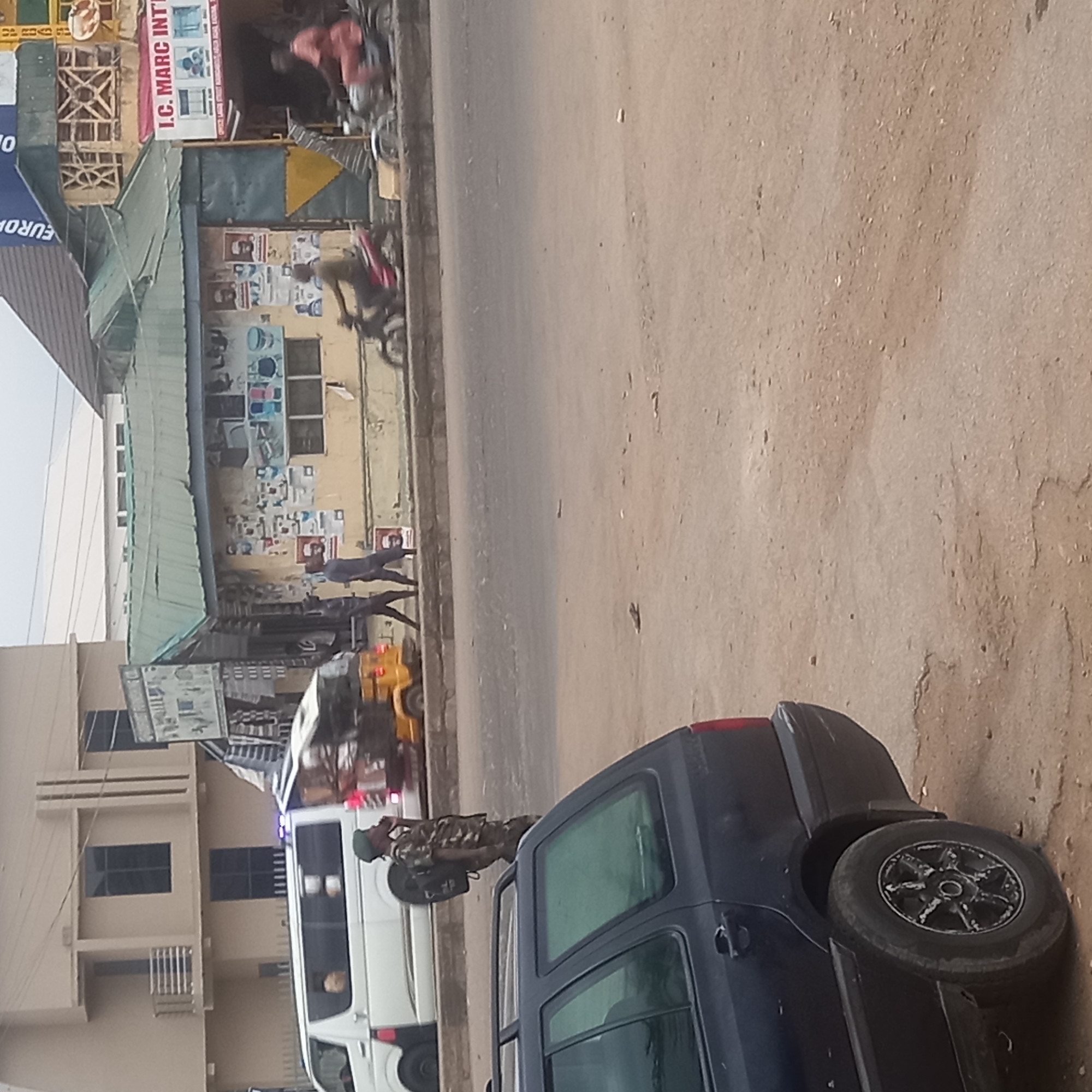 The image, when viewed upright, captures a daytime street scene in what appears to be an urban or semi-urban area, likely in Sardauna, Nigeria.
In the foreground, on the right side of the frame, a dark blue SUV with a spare tire mounted on its rear is prominently visible, parked or stationary on a dirt shoulder next to a paved road. Standing next to this SUV is a person dressed in a camouflage military-style uniform and a green beret, facing away from the camera.
Further along the road, a white minibus, possibly a commercial passenger vehicle or an emergency service vehicle due to blue flashing lights on its roof, is also present. A yellow motorized tricycle, commonly referred to as a "Keke Napep" in Nigeria, is visible beyond the minibus. Several motorcycles are seen parked or moving in the vicinity.
To the left of the road, a commercial building with a light green awning serves as a focal point in the middle ground. Its exterior wall is extensively covered with numerous posters and advertisements, some featuring images of people and various household items. A sign on this building clearly displays the text "I.C. MARC INT'L". Another partial sign to its left shows "EUROPA" and "O". Behind this shop, other multi-story buildings with windows line the street.
The scene depicts general street activity with people walking on the sidewalk near the shops. The weather is clear and sunny, suggesting a bright day. The overall atmosphere is that of a busy street in an active community.