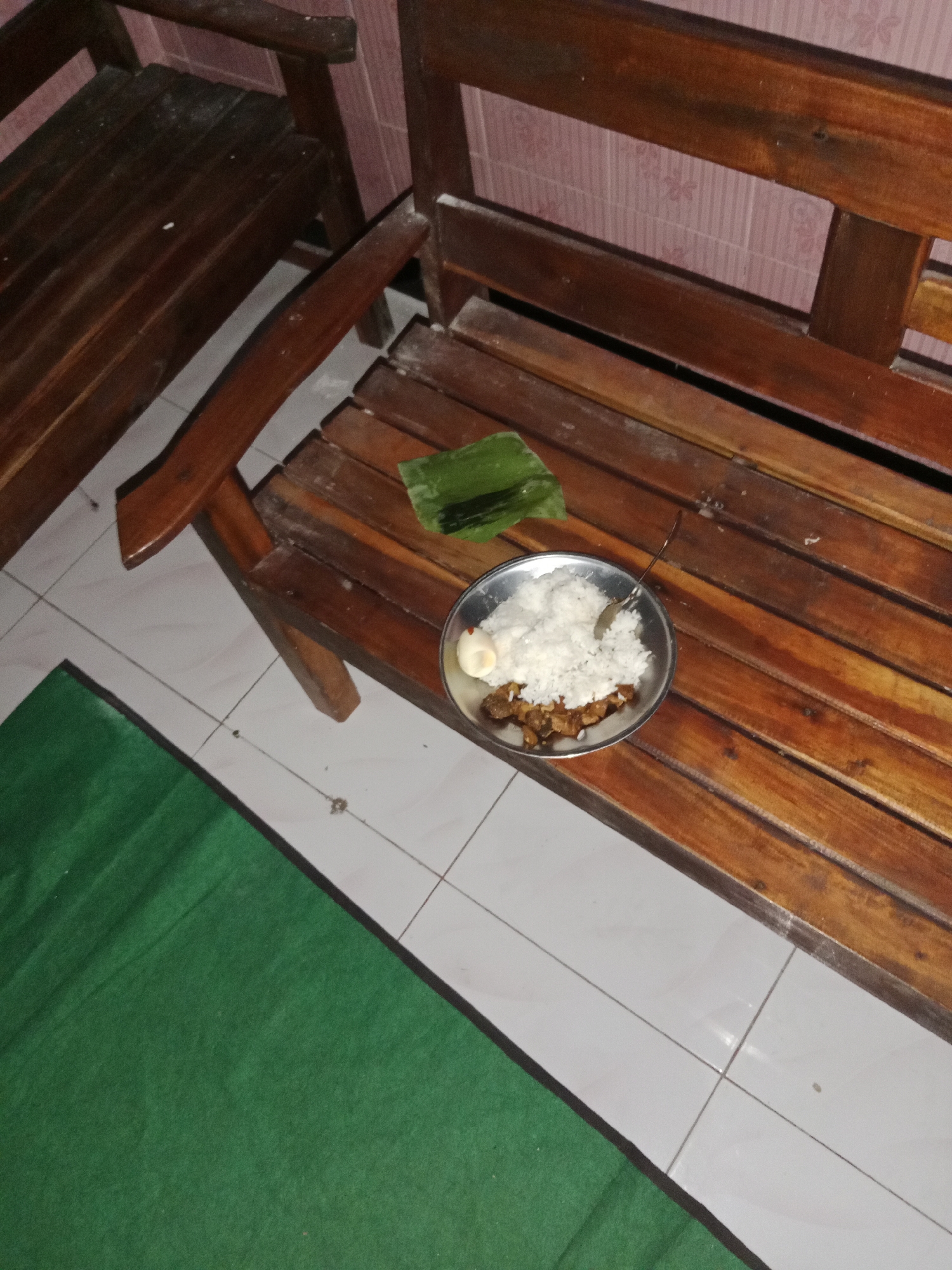 An overhead shot shows a meal of rice, a hard-boiled egg, and meat served on a metal plate, resting on a wooden bench in Ringinrejo, Indonesia. A green banana leaf is placed beside the plate, and a fork is sticking out of the rice. The bench is made of dark wood with visible grain and appears weathered. The floor beneath the bench is tiled in white with thin gray grout lines. To the left of the bench, a section of a green mat is visible. The background wall is tiled with a subtle pink pattern. The lighting is dim, suggesting an indoor setting.
