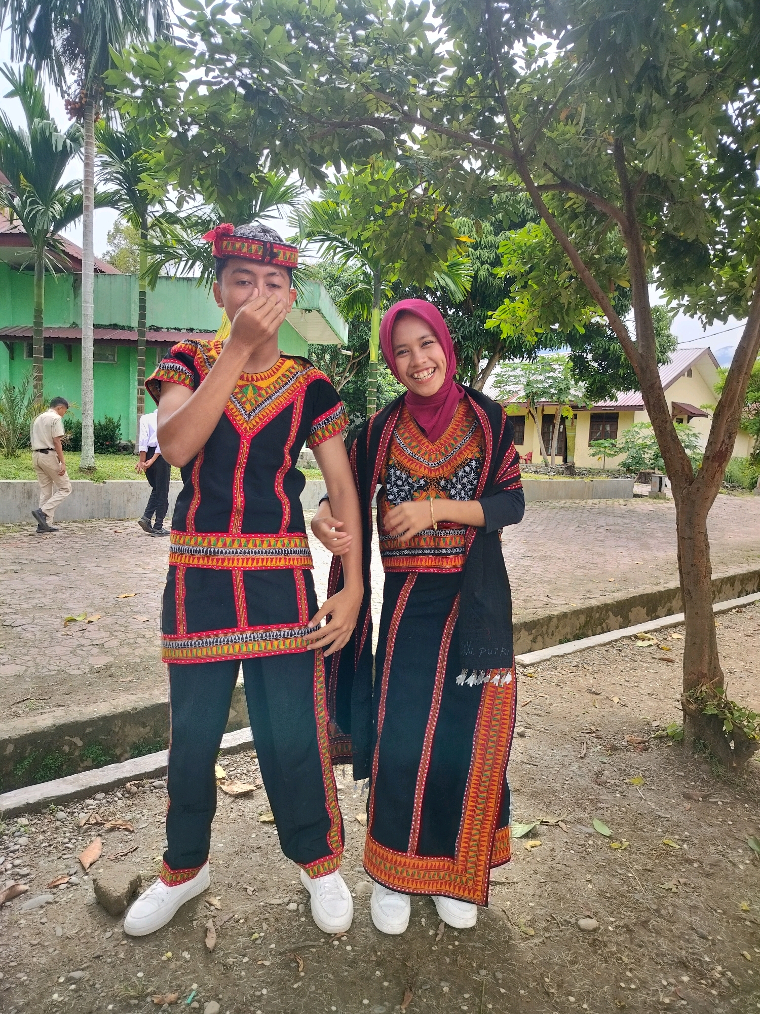 This media file features two young people dressed in traditional attire, posing for a photo outdoors. The individual on the left appears to be a male, wearing a black tunic with intricate colorful embroidery around the neckline, sleeves, and waist, paired with black pants and white sneakers. He has a red and black patterned headdress and is covering his mouth with his hand, smiling slightly. The individual on the right is a female wearing a matching black tunic and skirt, also adorned with colorful patterns, and a deep red hijab. She is smiling broadly and wears a gold bracelet on her left wrist. She also has a black shawl draped over her shoulders, which has embroidered patterns and tassels at the hem.
The scene is set in an outdoor area that appears to be a school or community ground, with a green building in the background, along with a light yellow building with a red roof. There are several trees with lush green foliage, suggesting it is daytime. The ground is covered with gravel and fallen leaves, and a concrete border runs along the foreground. In the background, two other individuals are walking away from the camera, one in a light-colored shirt and pants, and the other in darker clothing. The overall atmosphere is casual and cheerful, with the subjects exhibiting positive emotions. The image was taken in Purwodadi, Indonesia. The skirt of the female subject has visible text embroidered on it that reads "AN. PUTRI".