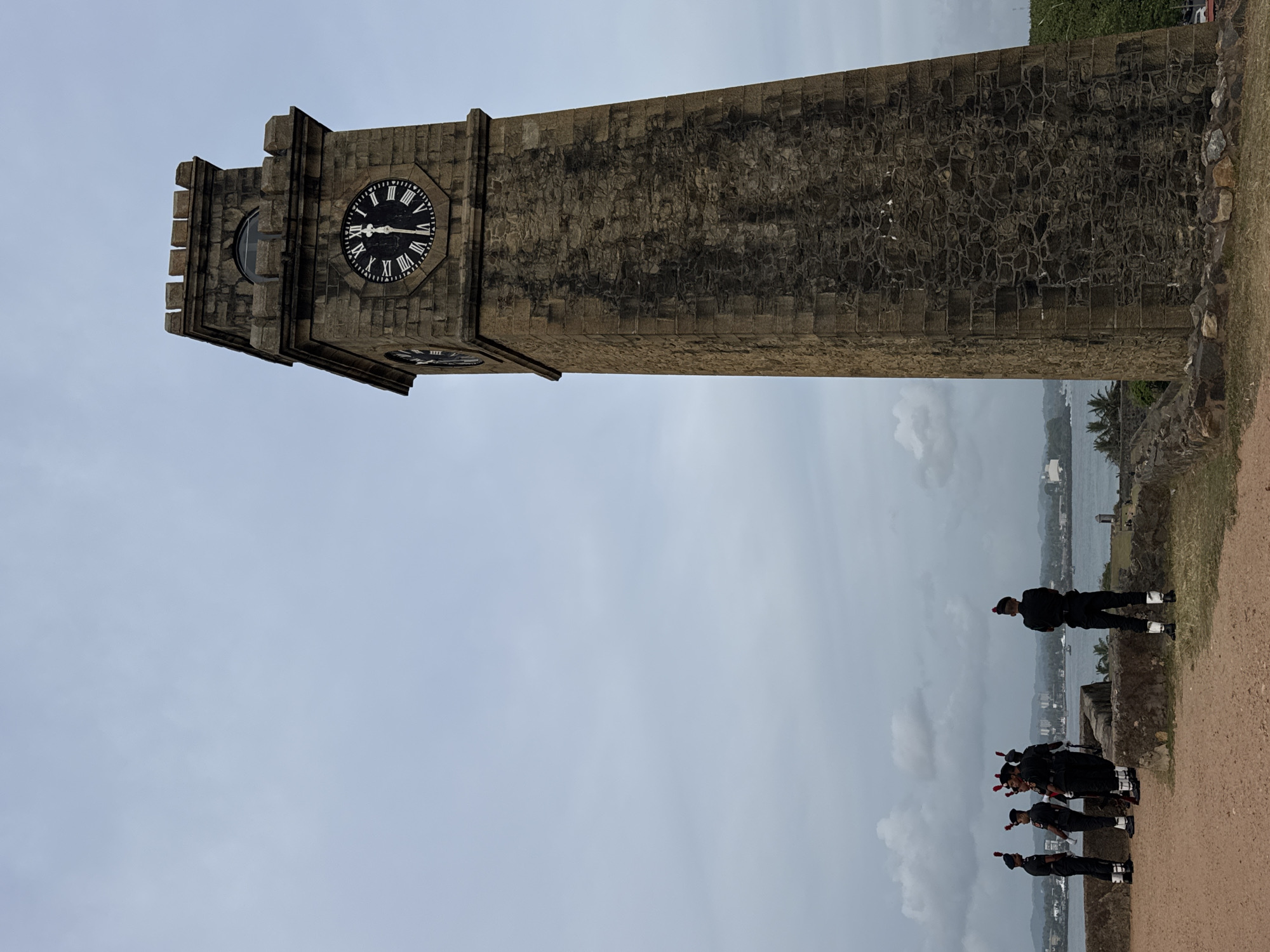 This is a full, eye-level shot of the Galle Fort Clock Tower in Galle, Sri Lanka, under a cloudy sky. The tower, a prominent landmark, is constructed of stone blocks and features a clock face with Roman numerals, indicating the time is approximately 10:30. To the left of the tower, a group of five soldiers in uniform stand on a dusty, elevated ground overlooking the sea. The soldiers are dressed in dark uniforms with red accents on their hats. In the background, the sea is visible, with a distant shoreline dotted with buildings and palm trees. The foreground shows the texture of the ground and some low stone walls.