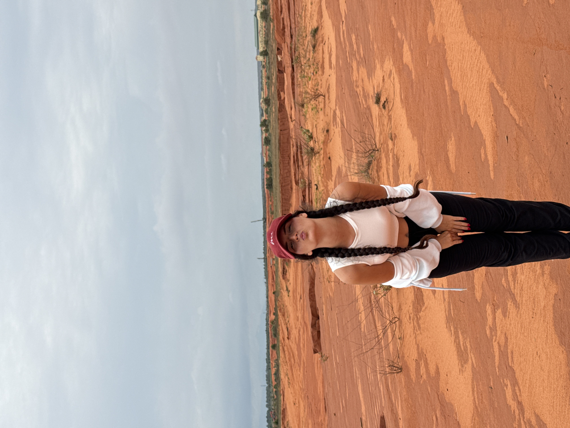 The image shows a young woman with dark hair in two braids, wearing a red baseball cap, a white crop top, and black pants. She is standing in a desert-like landscape with undulating orange sand dunes and sparse vegetation. The sky is overcast. In the background, there are some distant buildings and structures, suggesting a more developed area beyond the natural landscape. The woman is making a kissing face towards the camera. This location is likely the Red Sand Dunes of Mui Ne, Vietnam, a popular tourist attraction known for its unique desert scenery.