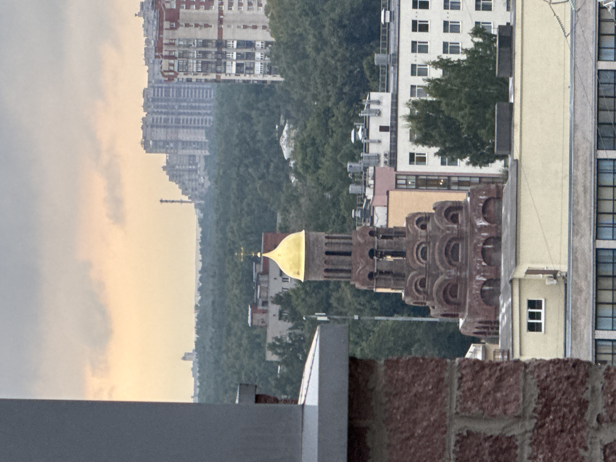 The image captures a city skyline with a prominent religious structure. In the foreground, a portion of a brick building is visible on the left, partially obscuring the view. A red brick tower with a golden dome and a cross stands out in the mid-ground, characterized by arched openings and intricate detailing. Behind the tower, a dense forest of green trees stretches across the landscape. Further in the background, several multi-story apartment buildings, characteristic of urban development, are clustered together, some with a stepped architectural design. The sky above is overcast with soft, muted colors, suggesting either dawn or dusk. The overall scene depicts a blend of religious architecture, natural elements, and urban sprawl in the city of Sinyavino, Russia.
