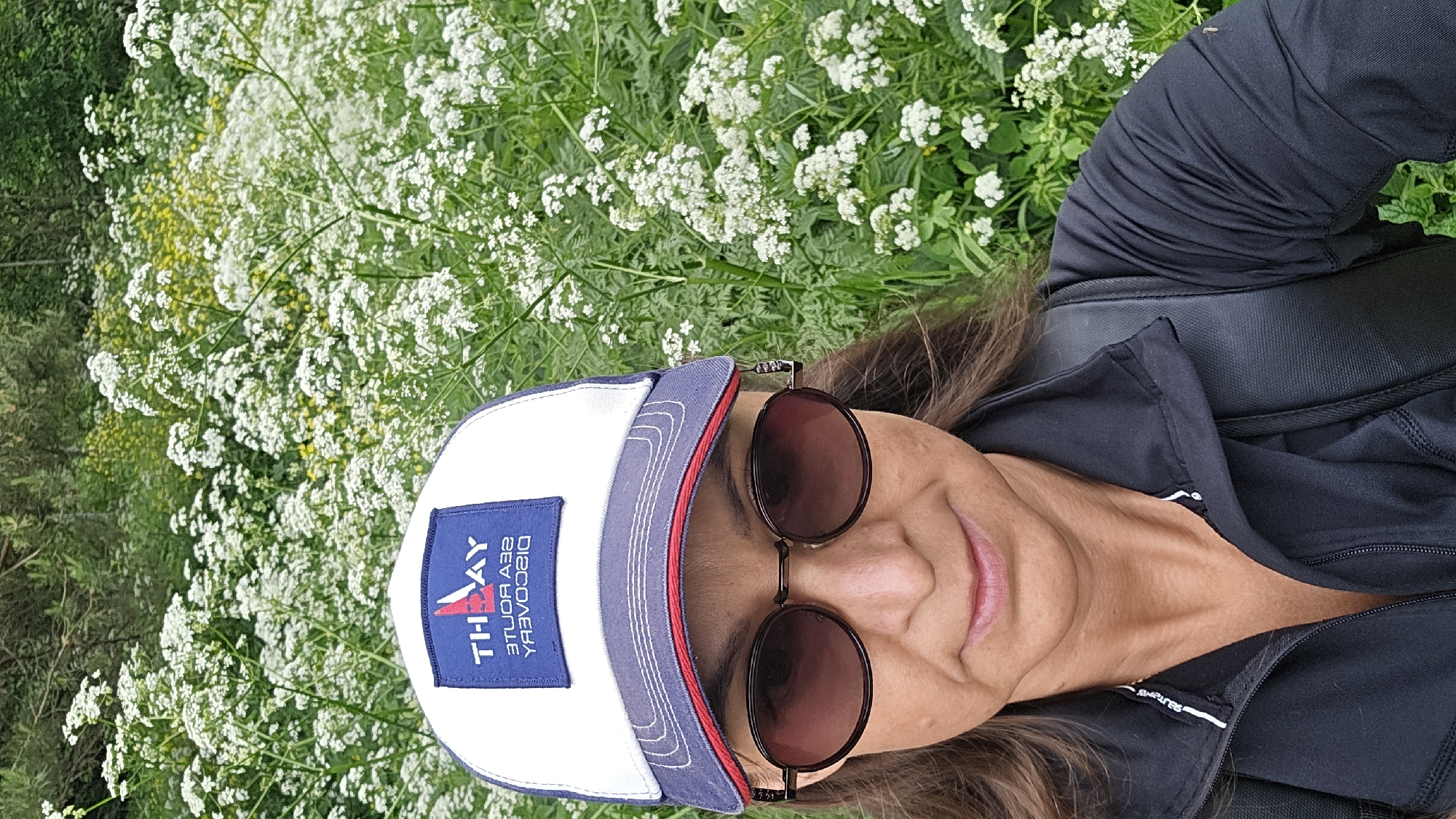 A woman with brown hair, wearing a black jacket, sunglasses, and a white and blue baseball cap, takes a selfie outdoors in front of a field of white flowers. The baseball cap has a logo that reads "THAY SEA ROUTE DISCOVERY" in blue and red lettering. The woman's facial expression is neutral. The background consists of lush green foliage with numerous small white flowers and some yellow flowers visible. The image appears to be taken during daylight hours. There is no indication of the city of Chaikovsky, Russia.