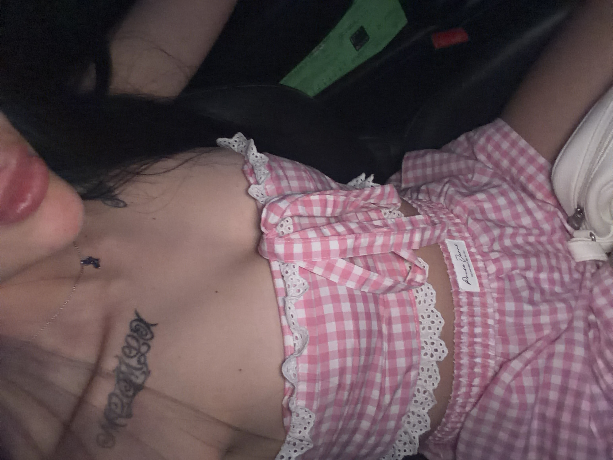 The image shows a close-up view of a person's chest, torso, and upper thighs. The person is wearing a pink and white checkered tube top with white lace trim and matching shorts. A tattoo is visible on the chest, and a delicate necklace with a small blue pendant is worn around the neck. The person's tongue is slightly visible. The background appears to be the interior of a car, with dark seating and a bright green object. There is no discernible indication of the location being Plovdiv, Bulgaria.
