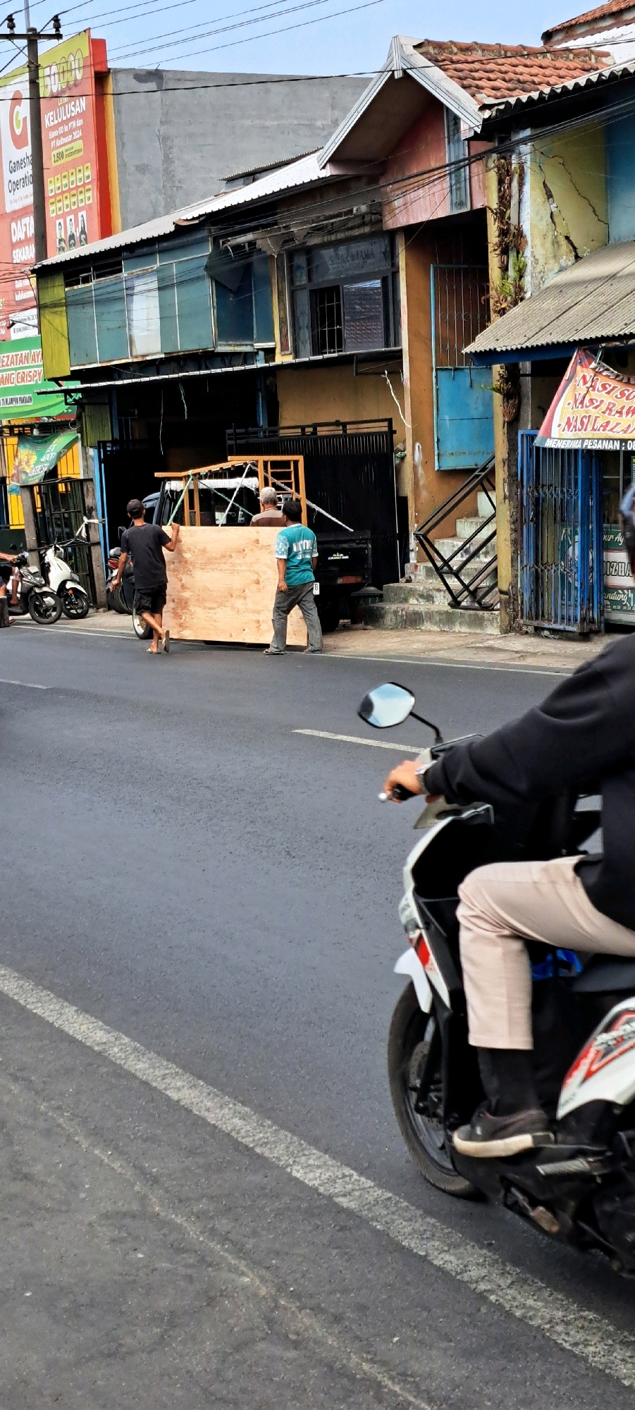In a street scene in Pandaan, Indonesia, two men are seen near the back of a dark pickup truck, one in a black t-shirt and shorts, the other in a teal t-shirt and grey pants. They appear to be moving a large, light-colored wooden panel. The pickup truck has a black metal frame structure at its rear. On the left side of the street, a white motorcycle is parked. In the foreground, the right side of a motorcycle, ridden by a person wearing a black sweatshirt and light-colored pants, is visible. The background shows commercial buildings with various signage, including "KELULUSAN" and advertisements for food. The architecture suggests a commercial or mixed-use area in a town. The road is asphalt with white lane markings. The sky is clear.
