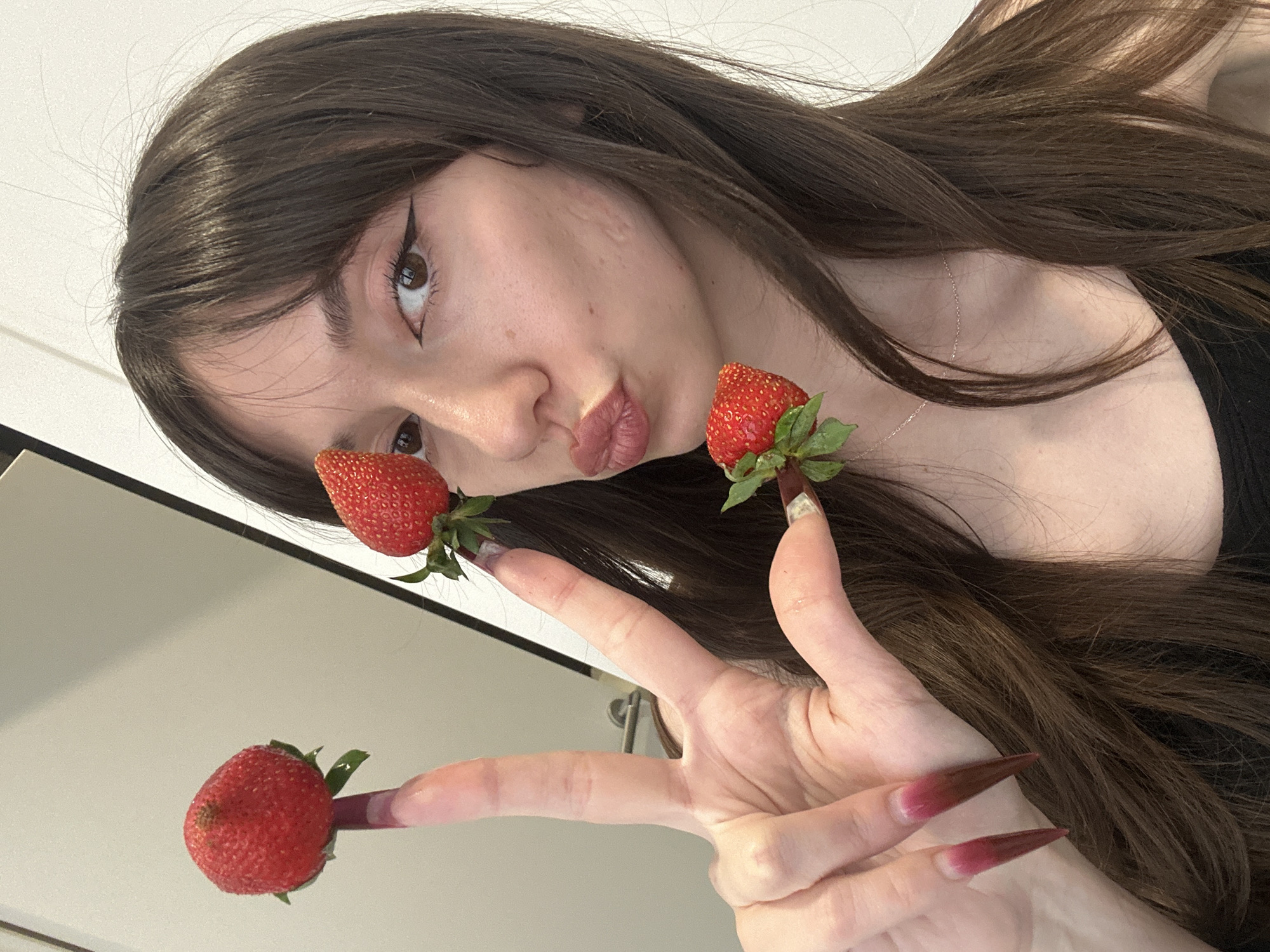 A young woman poses for a selfie indoors. She has long dark brown hair, brown eyes with eyeliner, and fair skin. She is wearing a black top and a delicate necklace. She holds three strawberries on her fingertips, one on her thumb, one on her index finger, and one on her middle finger. Her fingernails are long and painted with a gradient from pink to dark red. The background includes a white wall and a white door with a silver handle.
It's not possible to determine the specific location in the city of Vracar Urban Municipality, Serbia, from this image alone.