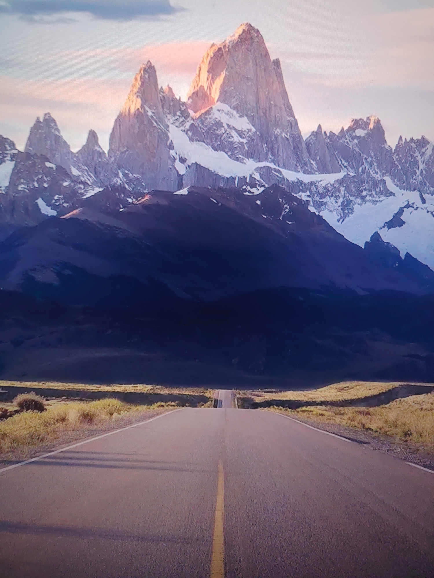 A road leads towards a mountain range with snow-capped peaks. The central peak is the tallest and catches the light. The mid-ground consists of a dark, hilly landscape. Low vegetation lines both sides of the road. A yellow line marks the center of the road. The sky is partially visible above the mountains.