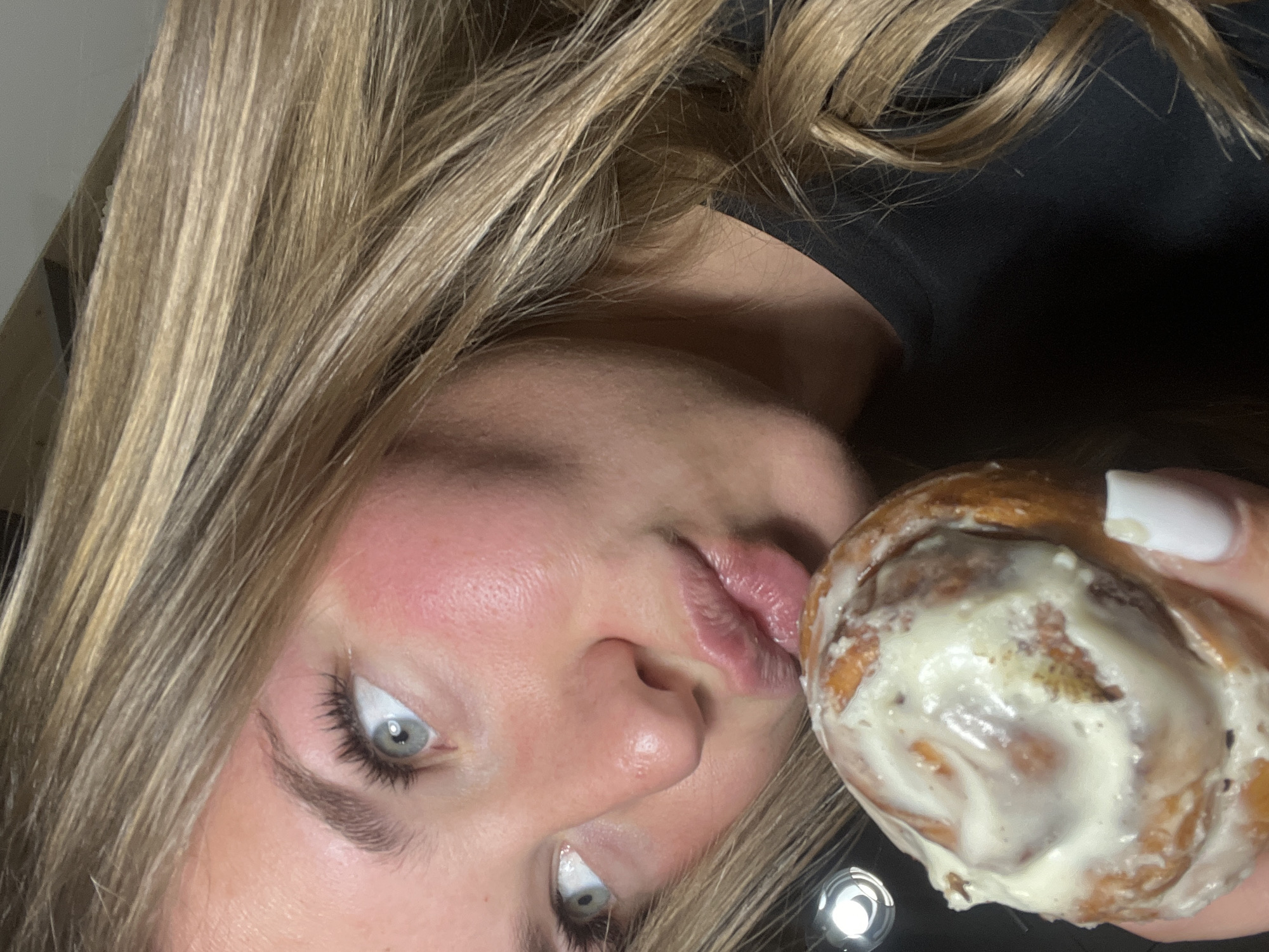 A blonde woman with light eyes is holding a frosted cinnamon roll close to her face. Her face is turned to the left, and her eyes are looking upwards. She is wearing a black shirt. The background is indistinct. The location of the photo is impossible to be determined from the image alone.