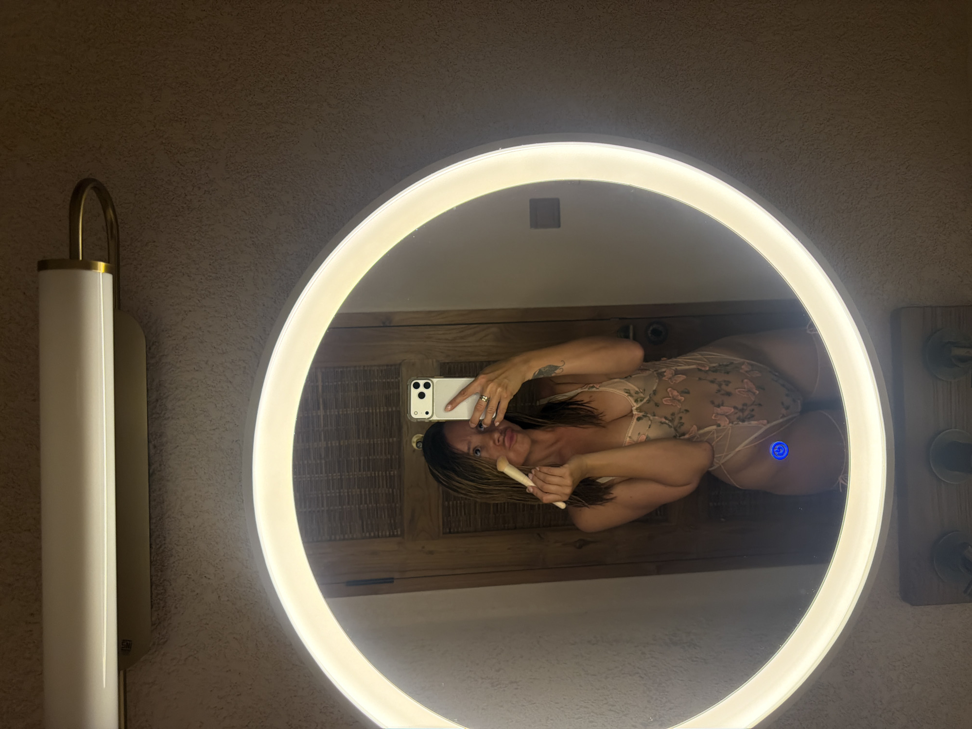 A woman is reflected in a large, circular, backlit mirror, taking a selfie. She is wearing a light-colored, sheer, floral-patterned lingerie set. She holds a makeup brush in her right hand and her phone in her left. Her expression is neutral, and she has long, dark hair with lighter streaks. There are visible tattoos on her arms.
The mirror is surrounded by a bright, white light that illuminates her and the background. The background visible in the mirror appears to be a room with wooden paneling and a woven texture. To the left of the mirror, a modern wall sconce with a white cylindrical shade and a brass accent is mounted on a textured wall. To the right of the mirror, there are some visible bathroom fixtures. The lighting suggests it could be evening or nighttime, or that the room is dimly lit naturally. There is no visible text in the image. The setting appears to be a bathroom or a dressing area in a hotel or residence, given the style of the mirror and the sconce. The overall mood is intimate and personal.