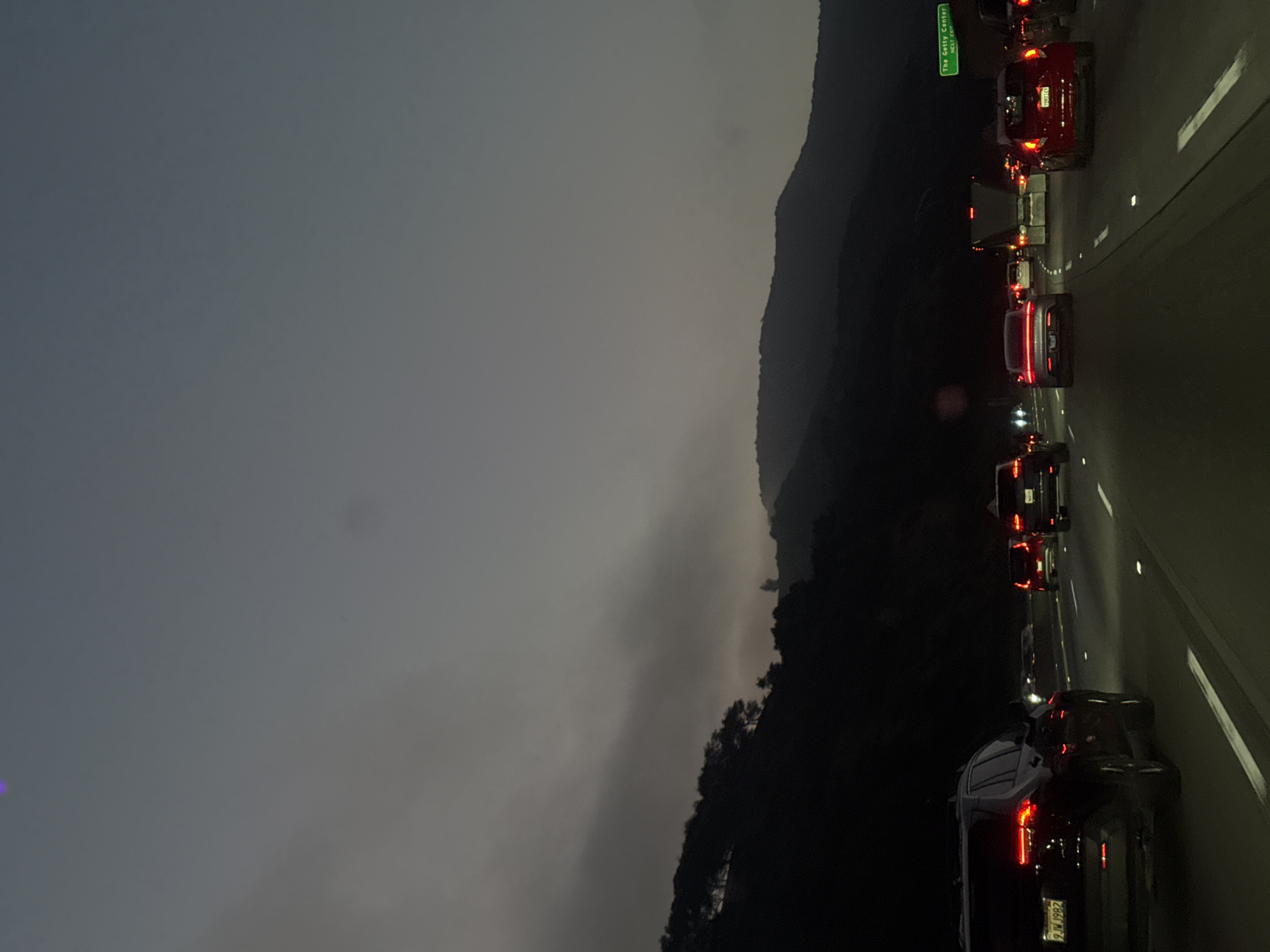 The image captures a multi-lane freeway scene at dusk or early night, characterized by heavy traffic and mountainous terrain. The location is the Interstate 405 freeway in Los Angeles, California, United States, identified by a green overhead sign on the right side of the road stating "The Getty Center NEXT EXIT".
The sky is dark gray, indicating low light conditions, with atmospheric haze or clouds partially obscuring the dark, tree-covered mountains bordering the freeway on both sides. The asphalt roadway features white dashed and solid lane markings, along with reflective road markers.
Multiple vehicles are present across several lanes, all traveling in the same direction. Their red taillights are illuminated, suggesting slow-moving or stopped traffic. In the foreground, on the far-left lane, a dark-colored SUV with California license plate "9JTW1982" is visible with its brake lights activated. To its immediate right, a gray sedan displays illuminated taillights. Further down the freeway, in the second lane from the right, a red hatchback with California license plate "39NRY14" is observed. A large semi-trailer truck is positioned in a central lane further ahead. Headlights from distant vehicles appear as bright white points of light. No individuals are visible outside of the vehicles. A faint purple light artifact is present in the upper left area of the sky.