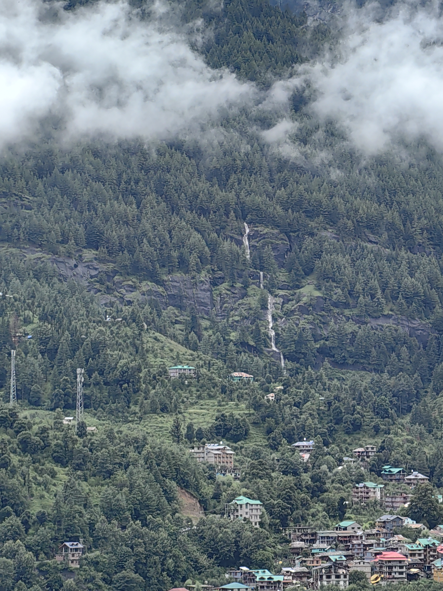 The image depicts a densely forested mountainside with a scattering of buildings and a waterfall. The location is identified as Vashisht, India. The upper portion of the image is partially obscured by white clouds that hang low over the trees.
The mountainside is covered in a thick growth of coniferous trees, appearing dark green in the subdued light. Several distinct waterfalls cascade down the rocky slopes, appearing as white streaks against the dark rock and trees.
Interspersed among the trees are numerous buildings, forming a village or town that sprawls across the lower and middle sections of the mountainside. The buildings vary in size and architectural style, with many featuring pitched roofs, some in green or other colors. There are also two prominent telecommunication towers visible on the left side of the image.
The overall impression is of a natural landscape with human settlement integrated into it, under a cloudy sky. No people are clearly visible in the image, and no specific actions or interactions are discernible. The scene appears to be static and focused on the landscape and the village.