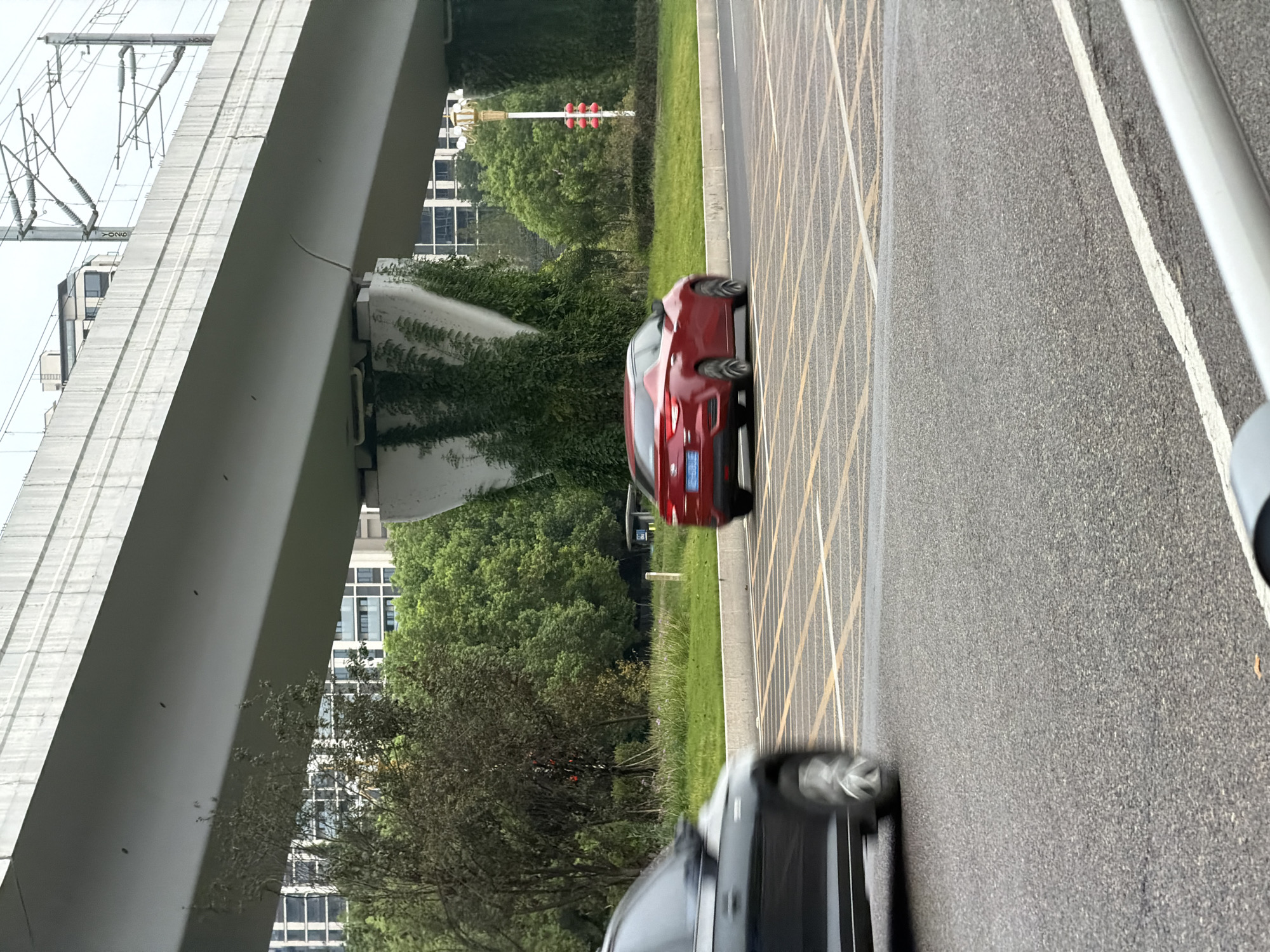 This image captures a street scene in Jiangjin, China. A red sedan is in motion on the road, transitioning across a set of yellow-lined road markings. To the left of the sedan, a blurred dark-colored vehicle is also moving. The background features lush green trees and modern buildings, suggesting an urban environment. An elevated structure, possibly a highway or a bridge, dominates the upper portion of the image, with some vegetation growing on its supports. The sky is overcast. The scene appears to be a typical day in a city with significant infrastructure and greenery.