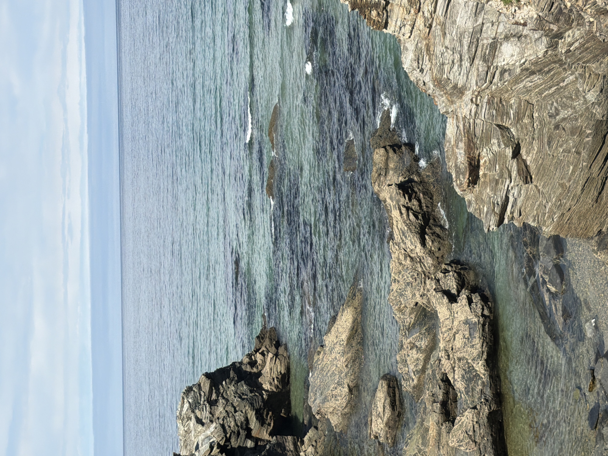The image captures a rugged coastline with clear, turquoise water lapping against layered rock formations. The sea extends to the horizon, meeting a sky with pale blue clouds. The rocks in the foreground display distinct horizontal striations, characteristic of sedimentary rock formations. Small waves gently break against the submerged and exposed rocks, creating white foam. The scene appears to be a natural landscape, devoid of human presence or artificial structures.
