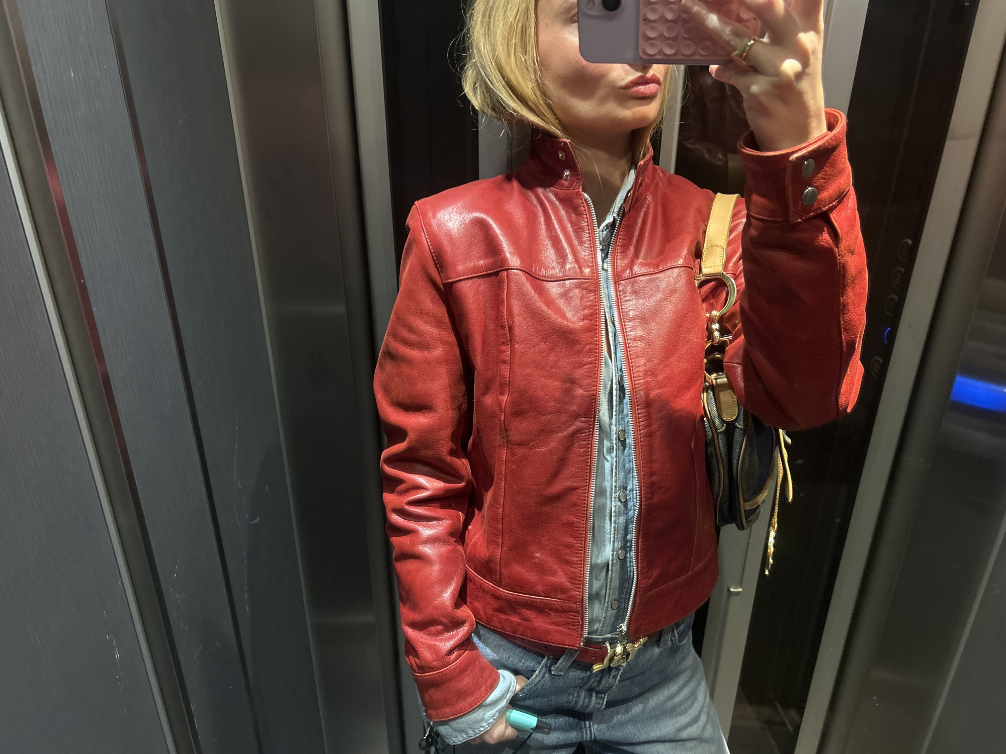 A person in blue jeans, a light blue button-down shirt, and a red leather jacket takes a selfie in an elevator. A handbag with a gold strap is draped over their shoulder. The elevator walls are gray with vertical lines. The city and specific location are unknown.