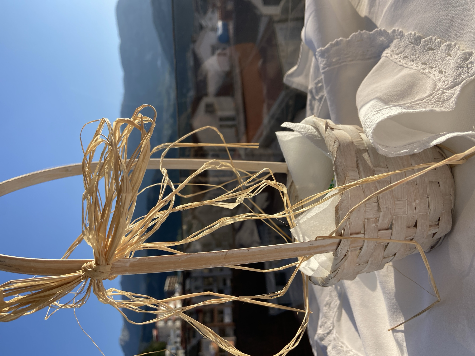 The image shows a small, woven basket with a handle adorned with raffia. The basket is filled with white paper napkins and is placed on a white tablecloth, possibly outdoors. In the background, a blurred view of a city landscape can be seen, featuring buildings and a mountain range under a clear blue sky. Based on the architectural style and mountain range, the location is likely Budva, Montenegro. There are no people visible in the image.