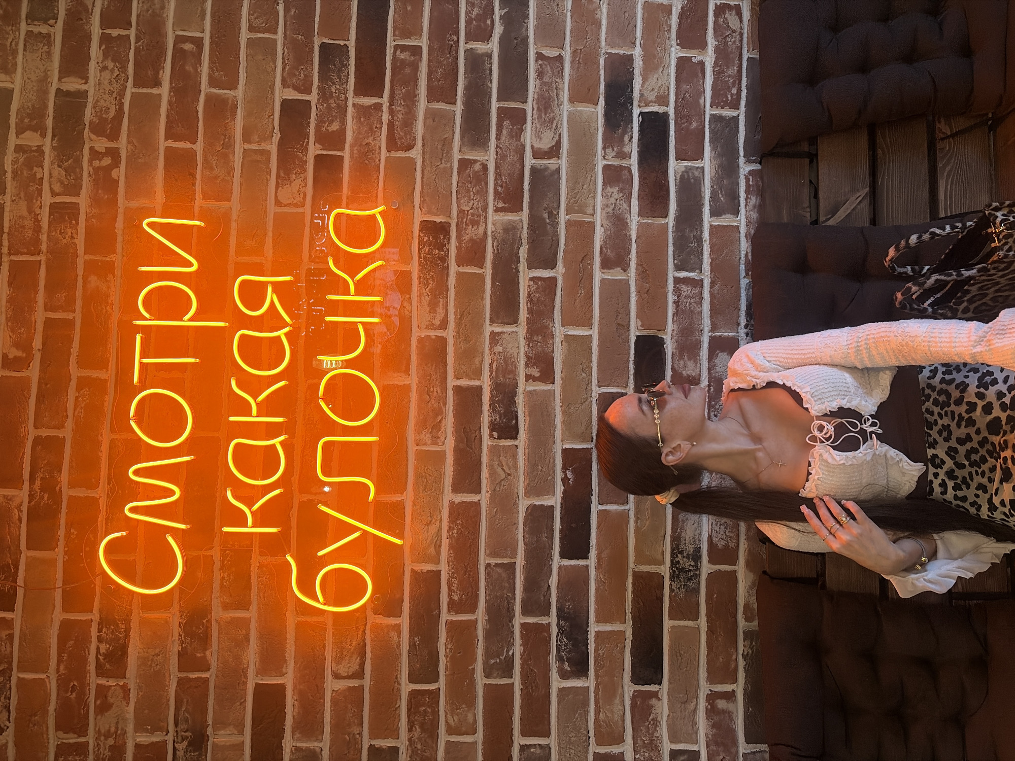An indoor scene depicts a female subject seated on a dark brown cushioned bench against a reddish-brown brick wall in an establishment located in Saint Petersburg, Russia. Above her, a bright yellow-orange neon sign displays the Cyrillic text 'Смотри какая булочка,' translating to 'Look what a bun!' or 'Look what a cutie!' The subject has dark hair pulled into a ponytail with a light scrunchie and wears gold-rimmed rectangular sunglasses. She is dressed in a white, textured, long-sleeved top with ruffles and a tie-front over a dark undergarment, paired with a leopard print skirt. A gold necklace with a cross pendant and several rings on her right hand are visible. She is positioned looking towards the right side of the frame, with her right hand resting on her chest. A leopard print bag is situated on the bench to her left.