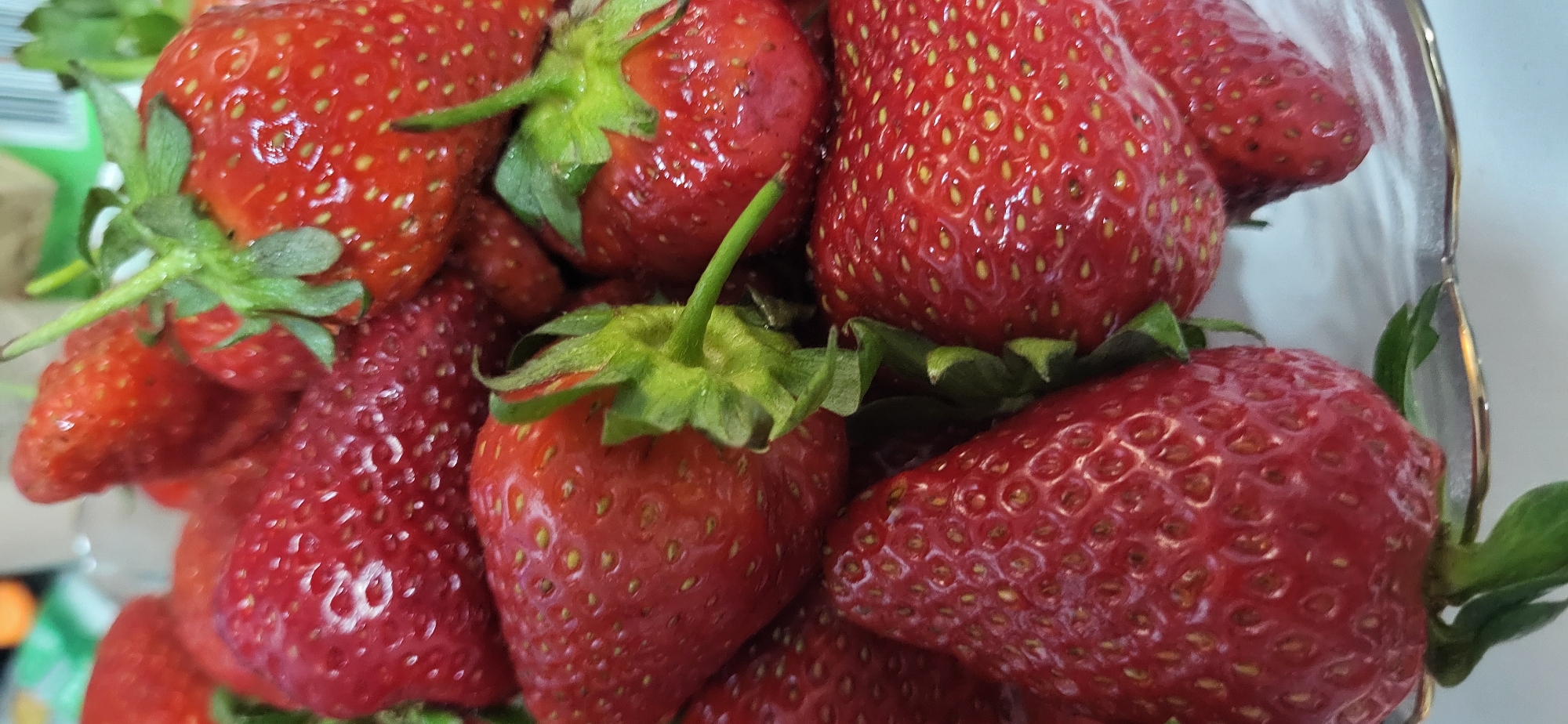 A close-up shows a cluster of fresh, ripe strawberries in a glass bowl. The strawberries are a deep red color with small yellow seeds dotting their surfaces. Green stems and leaves are attached to some of the berries. The bowl is partially visible, suggesting the strawberries are contained within it. The setting appears to be indoors, with a neutral background.
