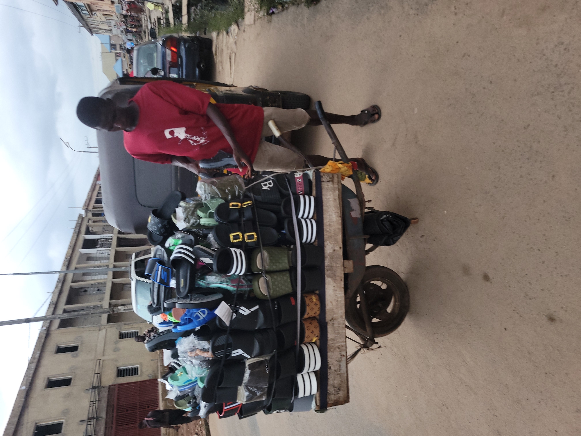 A man in a red t-shirt and light-colored shorts is pushing a cart filled with sandals. The cart is loaded with various styles of sandals, including flip-flops with stripes and sandals with decorative buckles. The man appears to be a vendor selling his wares.
The scene is set on a paved street in a commercial area, likely in Egbagu, Nigeria. Buildings line the street, with some appearing to be multi-story structures. A dark-colored vehicle is visible in the background, suggesting an urban or semi-urban environment. The sky is overcast, indicating a cloudy day.
The man's posture suggests he is actively engaged in moving his business. The overall atmosphere appears to be one of daily commerce. There is no discernible text visible in the image. The time of day is not explicitly stated but the lighting suggests it could be daytime.