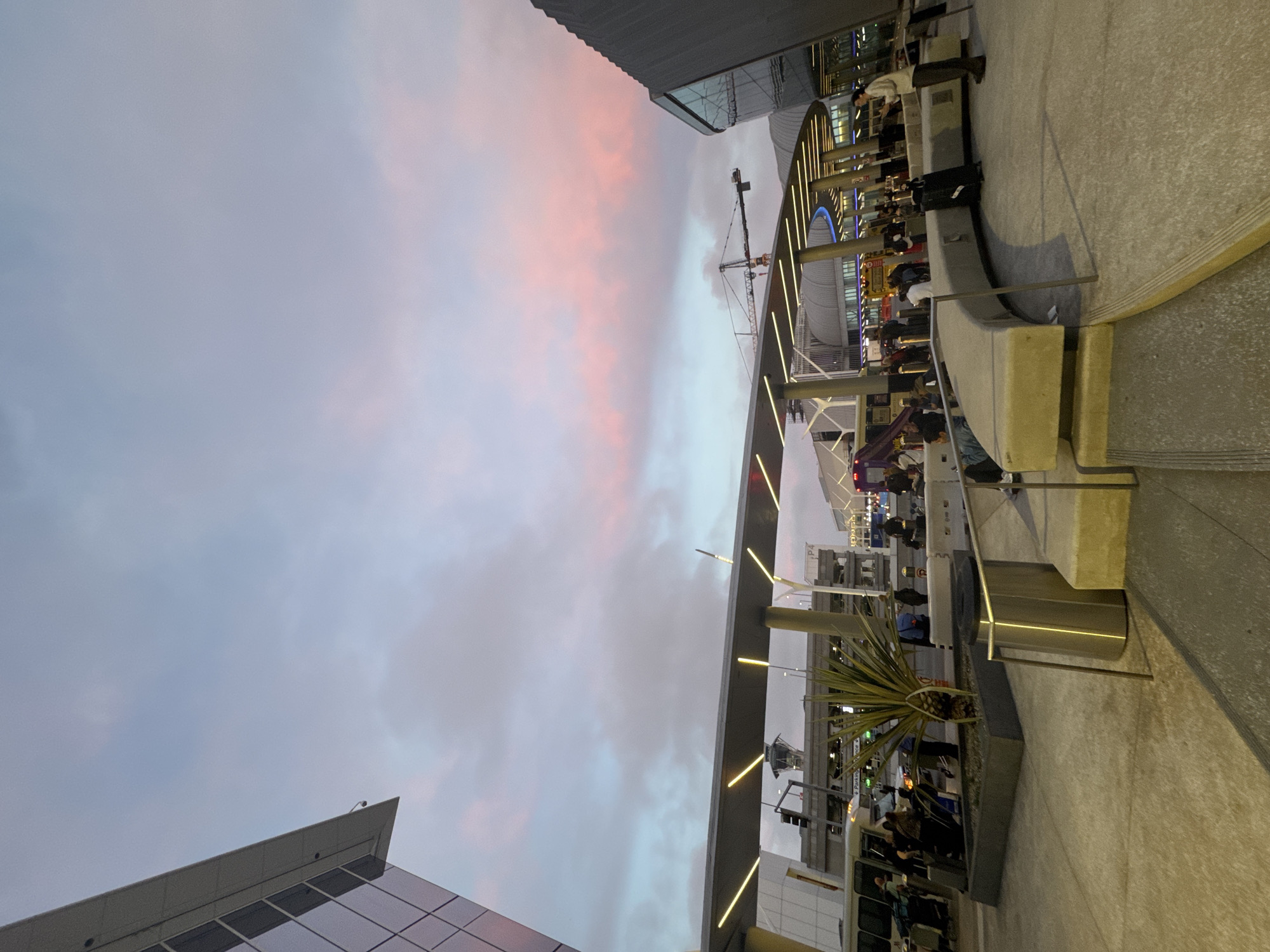 The image is a slightly tilted shot of a scene in Los Angeles, United States, likely at or near an airport. The sky is a prominent feature, with pink and grey clouds indicating either sunrise or sunset. Buildings are visible in the lower and upper left corners.
In the center of the image, an elevated walkway or structure with linear lighting runs diagonally. People are present, some standing in queues or walking, possibly waiting for transportation. There are plants in containers. A construction crane can be seen in the background.
The scene appears to be at an airport terminal or transportation hub with modern architecture and a bustling environment.