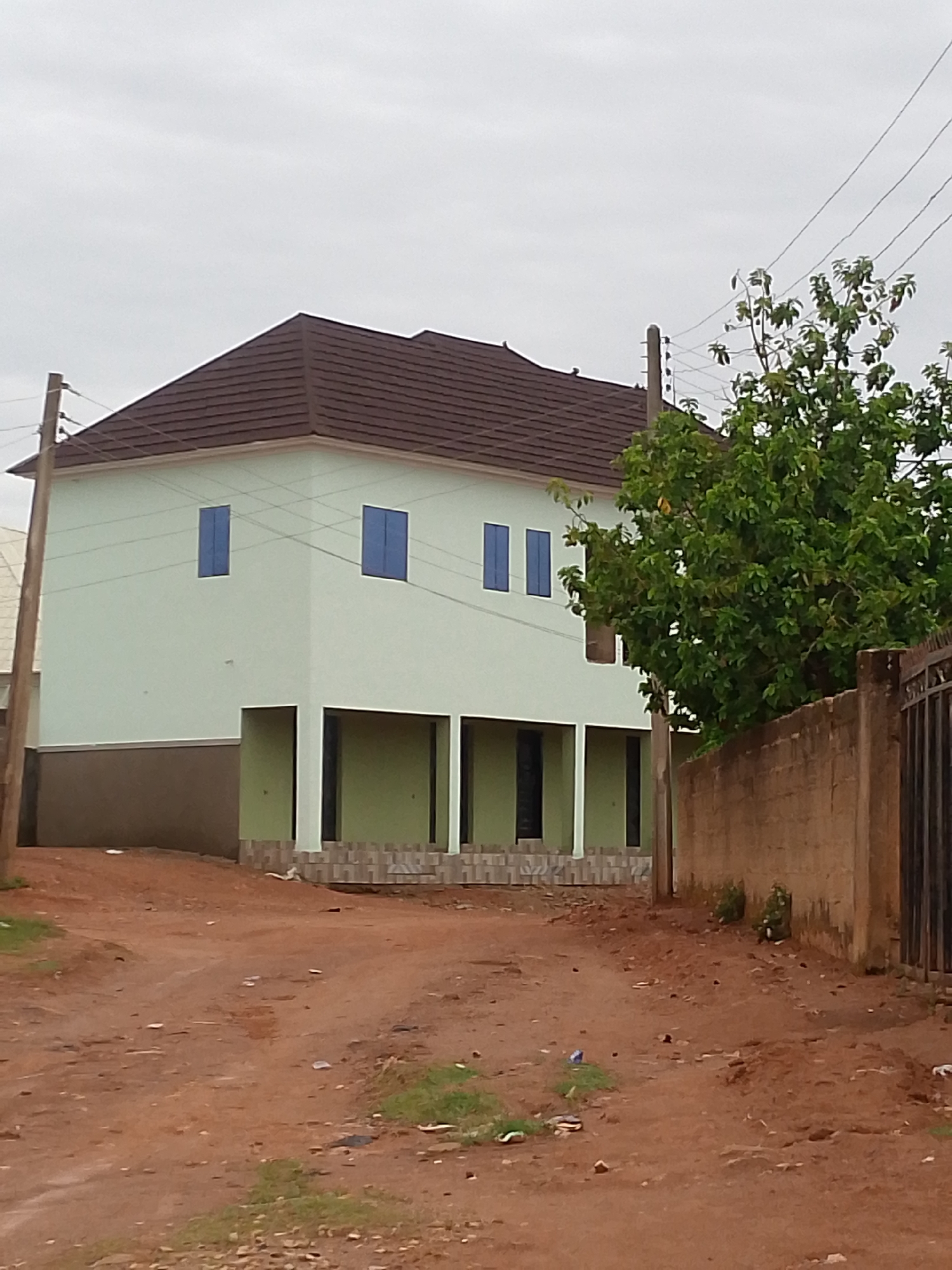 The image shows a two-story building in an urban area. The building is painted a light green color, with a brown roof and blue windowpanes. The ground floor has a series of dark green arches. The building sits at the intersection of a dirt road and a brown brick wall. A green tree sits behind the brick wall. A light brown wooden post sits in front of the building, running vertically from the ground to a brown electrical wire at the top of the frame. A dirty brown dirt road stretches across the bottom of the frame. The sky is overcast.