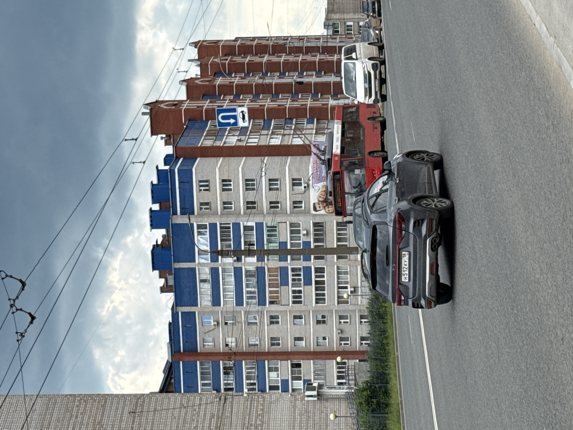 The image shows a street scene in Izhevsk, Russia. A dark gray car is in the foreground, traveling on a multi-lane road. Behind it, a red trolleybus and a white van are also on the road. In the background, there is a tall, modern apartment building with blue and gray facades, as well as a brick building with red brick accents. The sky is overcast. A traffic sign indicating a U-turn is visible on the right side of the road. The license plate of the dark gray car is visible and reads "H512KY 18". The overall scene depicts urban traffic on a cloudy day.