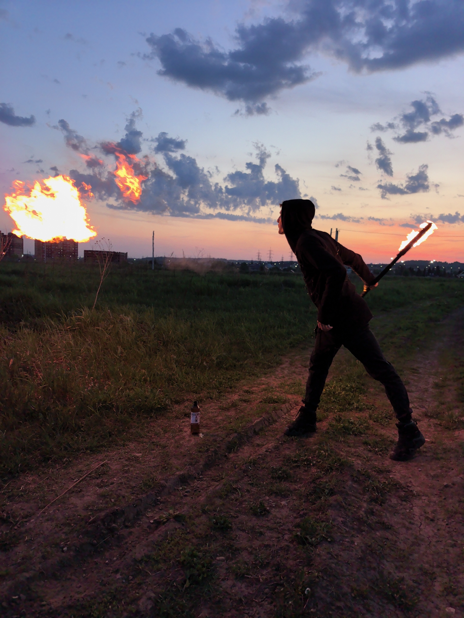 The image depicts an outdoor scene in a field, possibly near or within the city of Фролы, Russia. At sunset, a person wearing dark clothing, including a hooded jacket, stands in the foreground, holding a flaming object resembling a torch or staff in their right hand. The person appears to be positioned on a dirt path or ground, facing left. In the background, there is a controlled fire or explosion taking place, with a large plume of flames. A bottle, possibly a beer bottle, is visible on the ground near the foreground. Beyond the field and fire, the landscape includes a horizon with buildings and infrastructure, typical of a suburban or semi-urban environment. The sky displays a colorful gradient of sunset hues with scattered clouds.